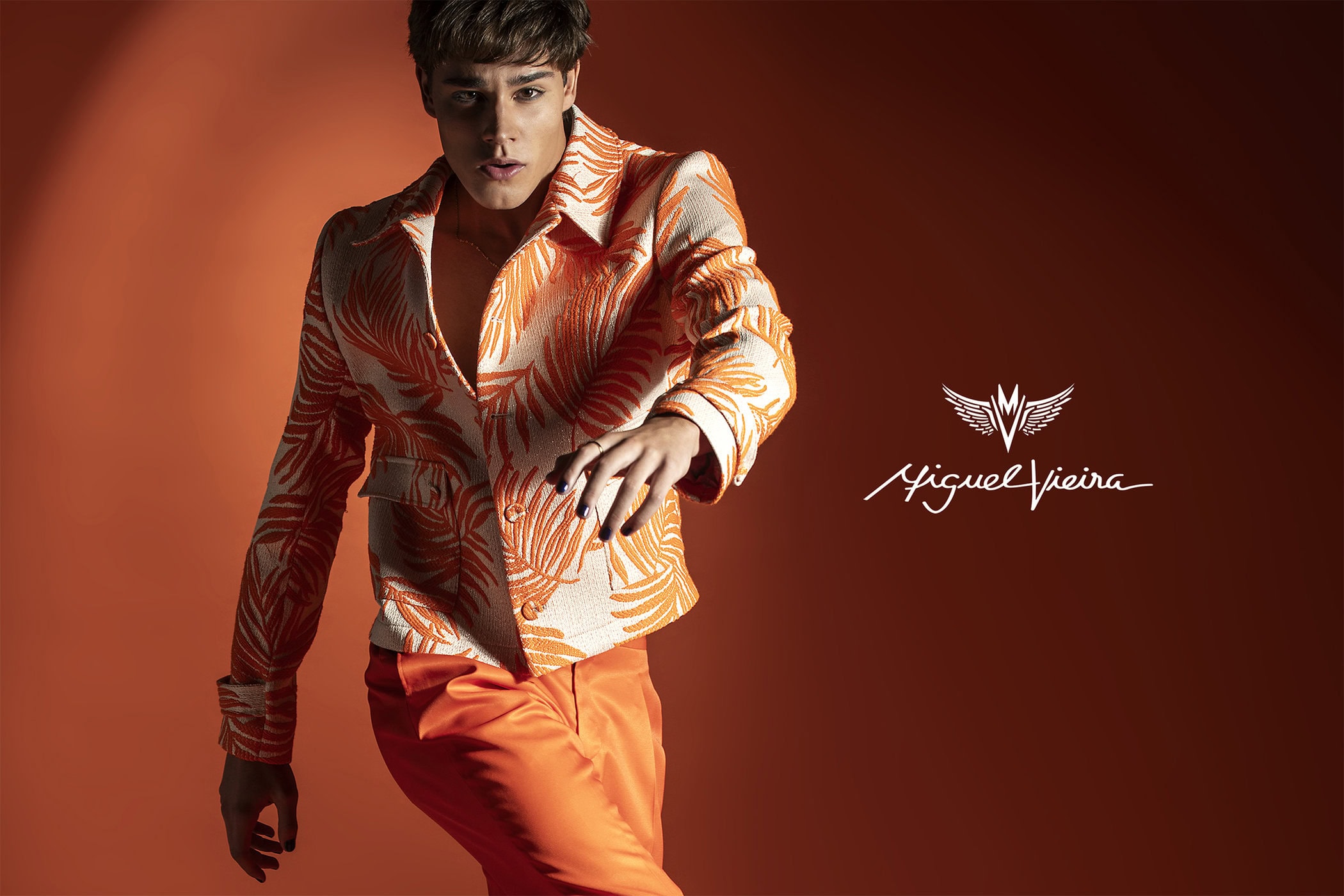 A young adult poses in an orange patterned jacket and matching pants against a solid orange backdrop in a fashion-forward studio portrait.