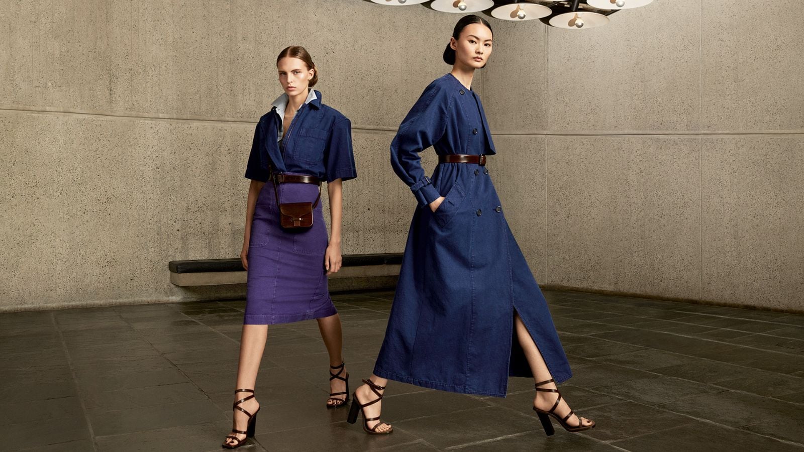 Two fashion models pose indoors wearing denim and purple skirts with strappy heels in a studio with a concrete background.