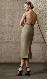 Model wearing a backless olive dress and strappy heels posing against a textured concrete wall for Max Mara