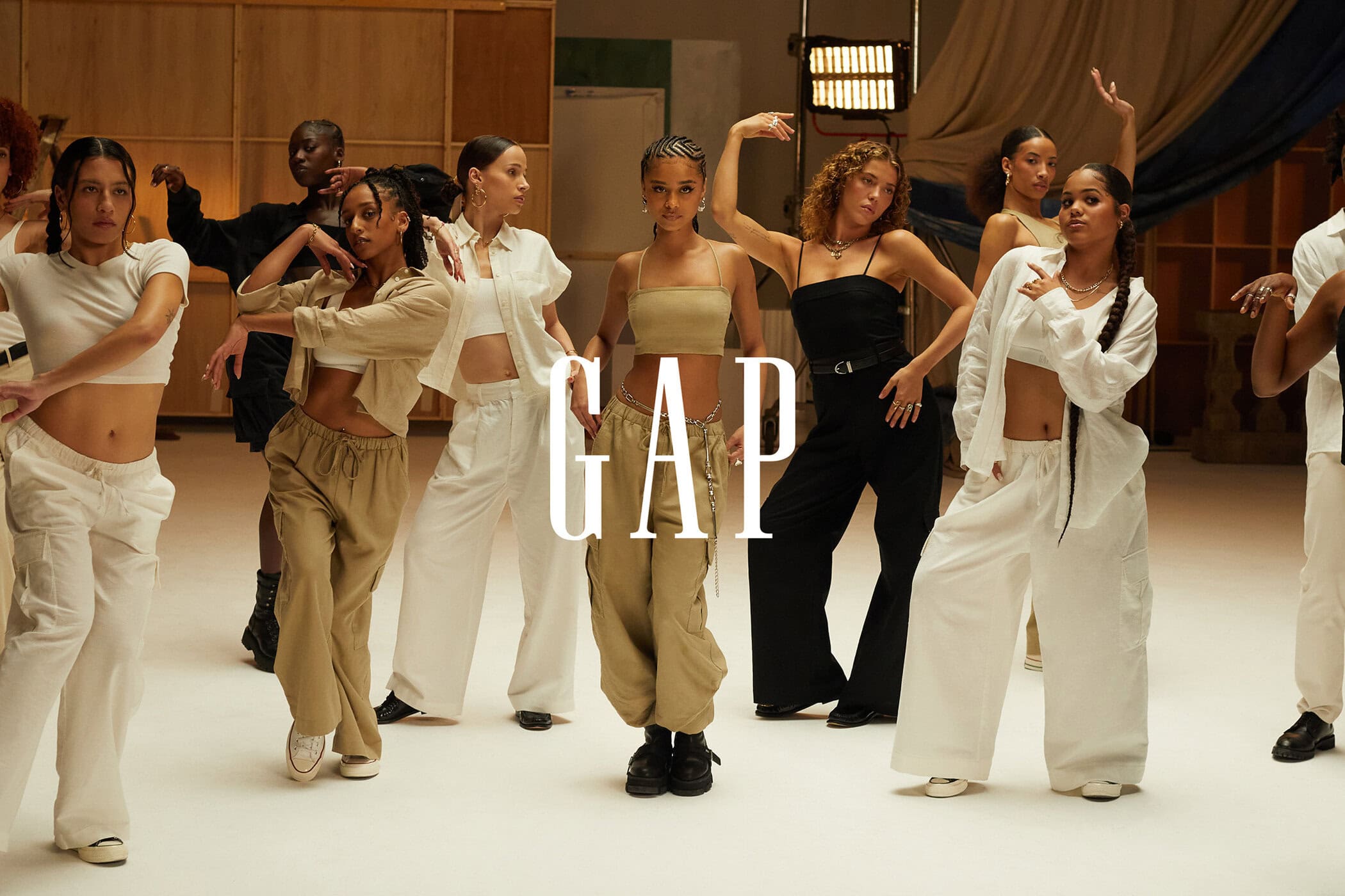 Group of young adult models wearing casual, neutral-toned Gap clothing perform a dance routine in a bright studio setting. 