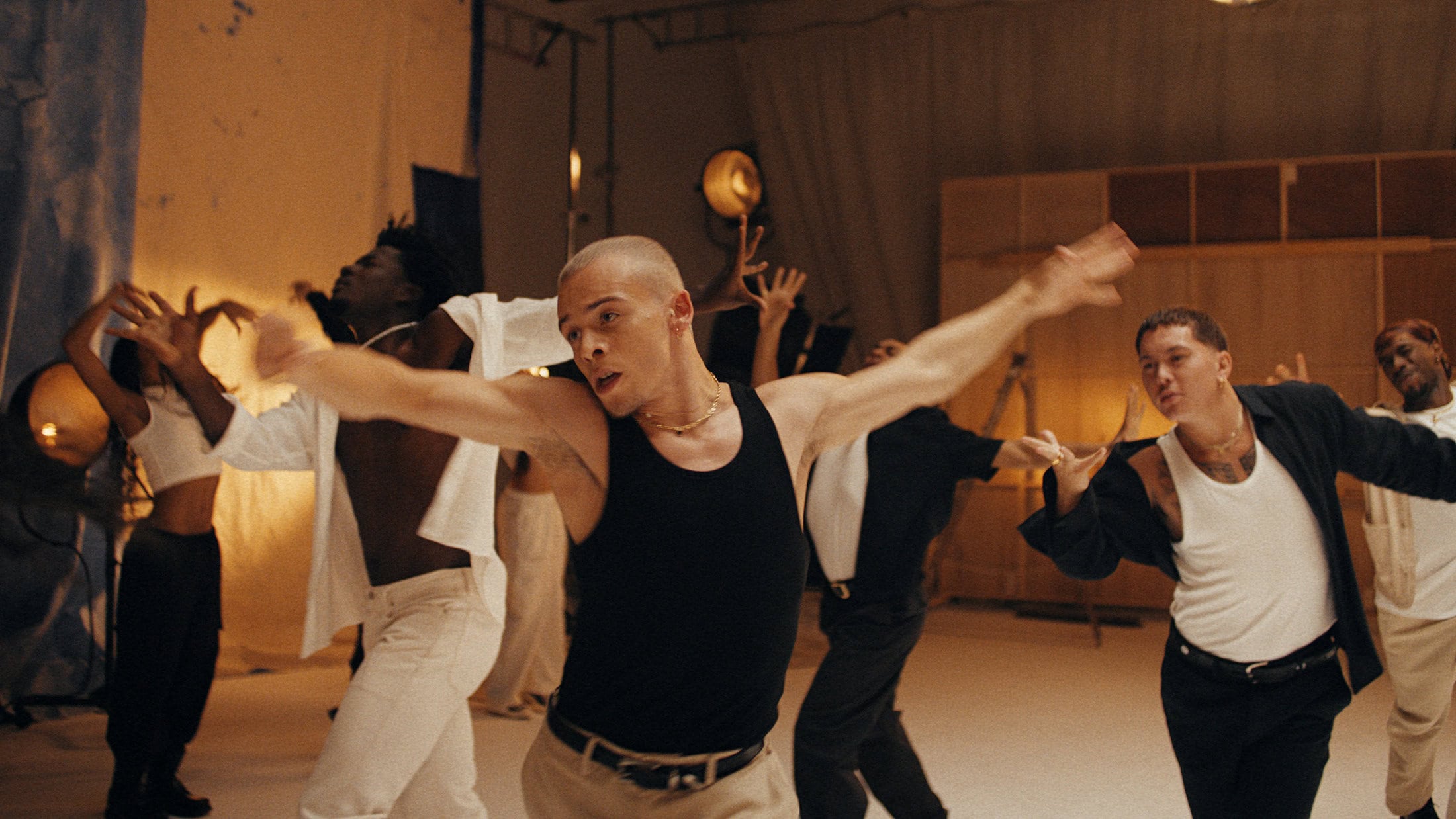 Group of diverse young adults dancing in a studio with warm lighting, wearing casual clothing and gold jewelry, and posing with arms raised.