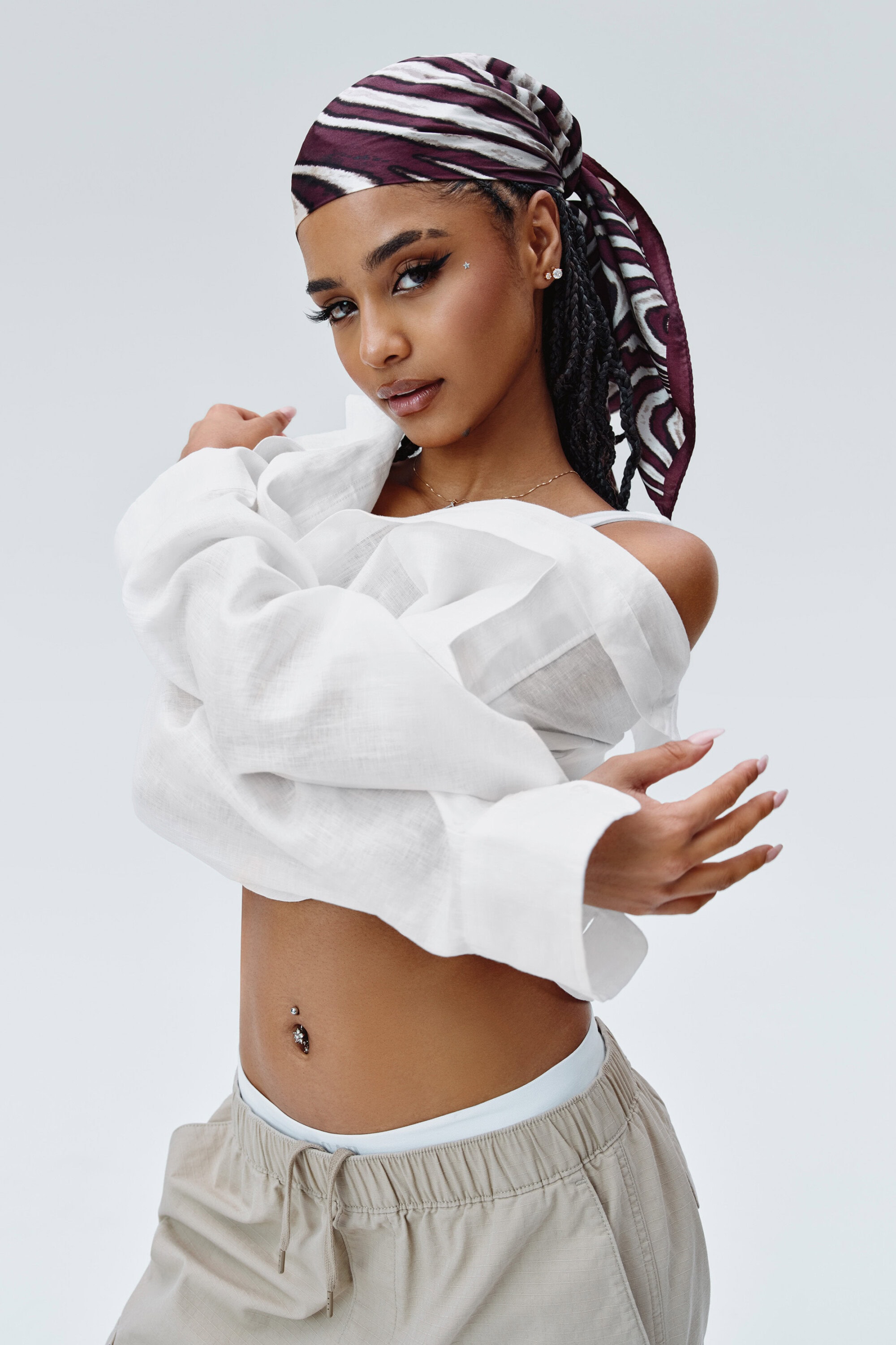 Young woman with headscarf poses in a white, off-the-shoulder shirt and neutral pants against a light backdrop in a fashion photo.