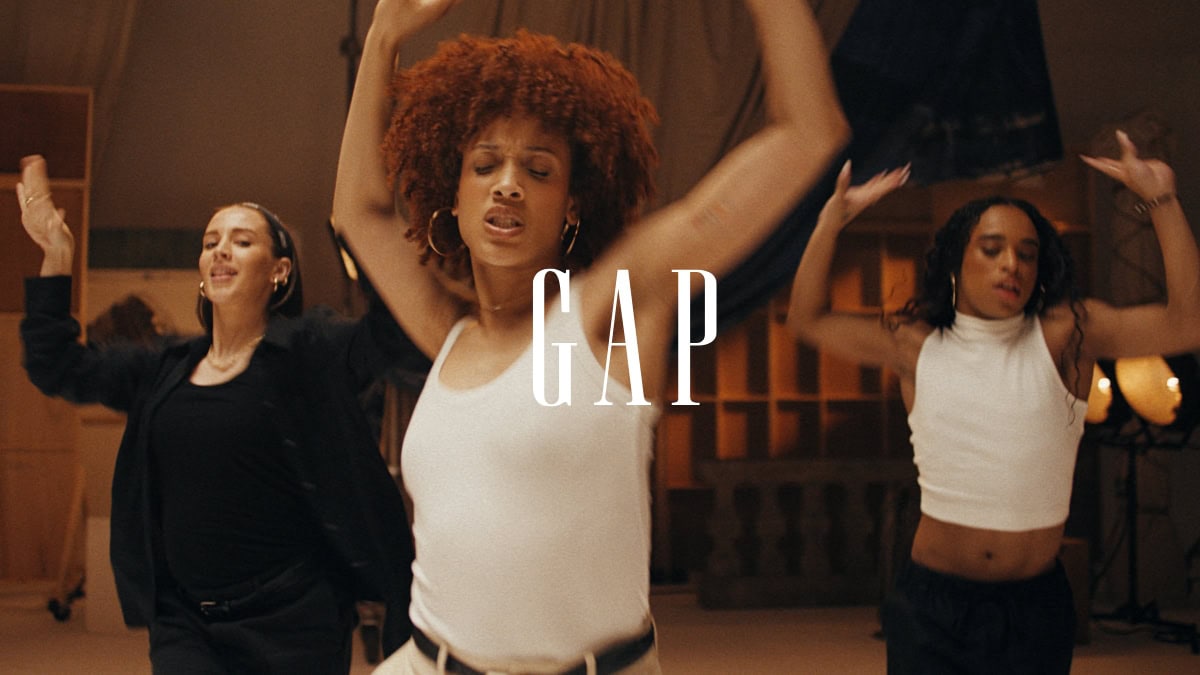 Three young adults are dancing and smiling in a bright photo studio for a GAP advertisement. The style is natural and vibrant.