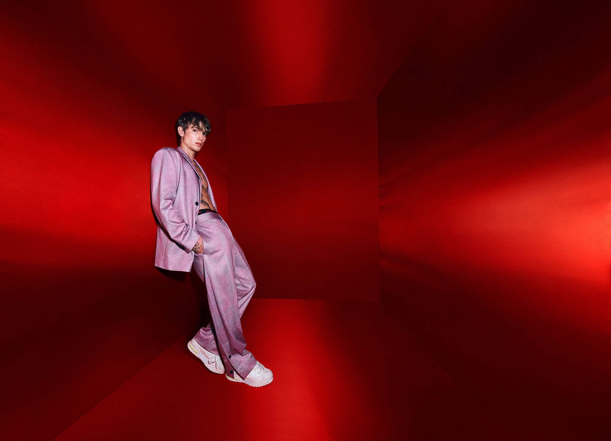 Young adult in a lilac suit leans against a red wall in a brightly lit photo studio, posing confidently and looking at the camera.