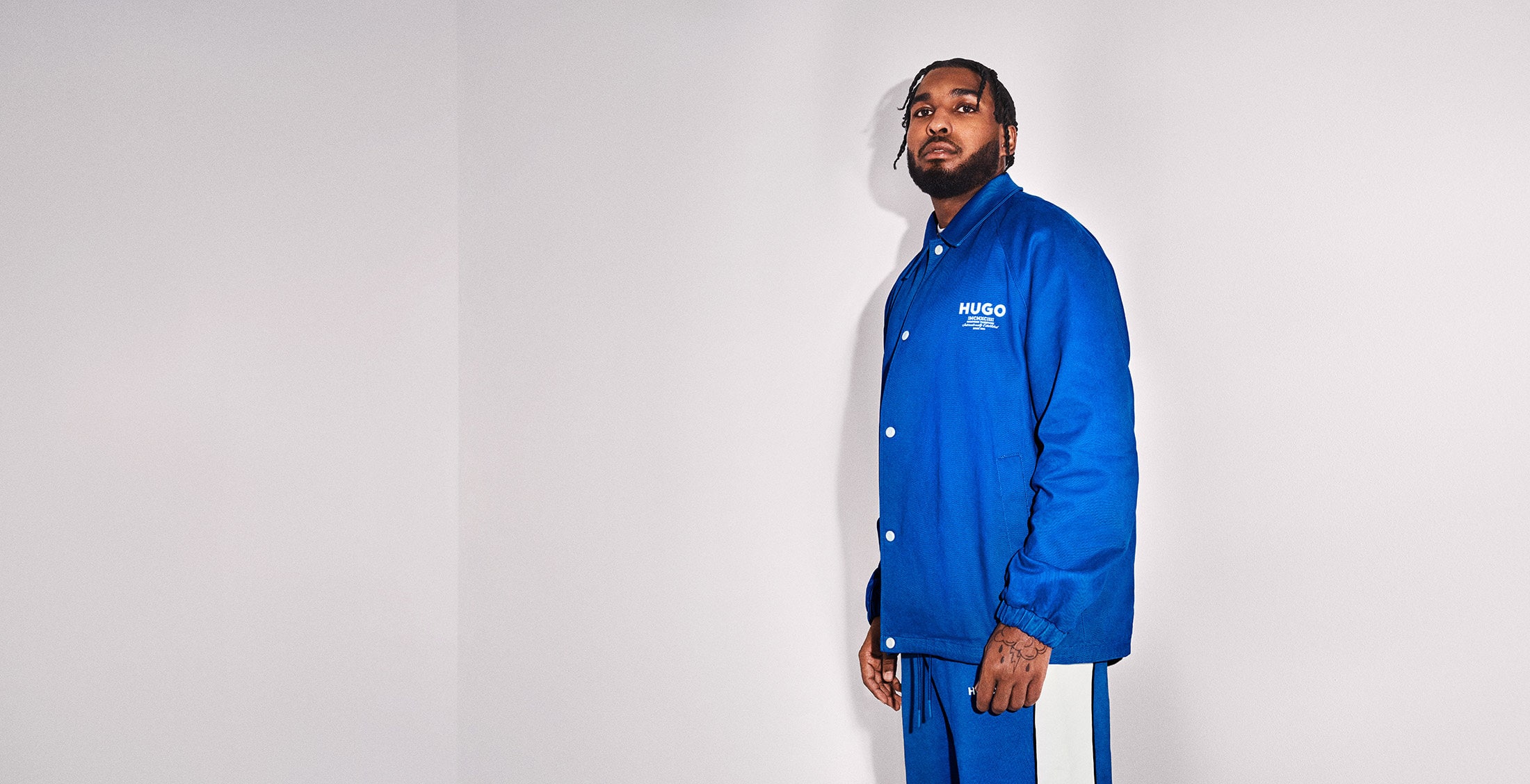 Adult male wearing blue jacket and matching blue pants poses in front of a white wall in a studio with soft lighting.