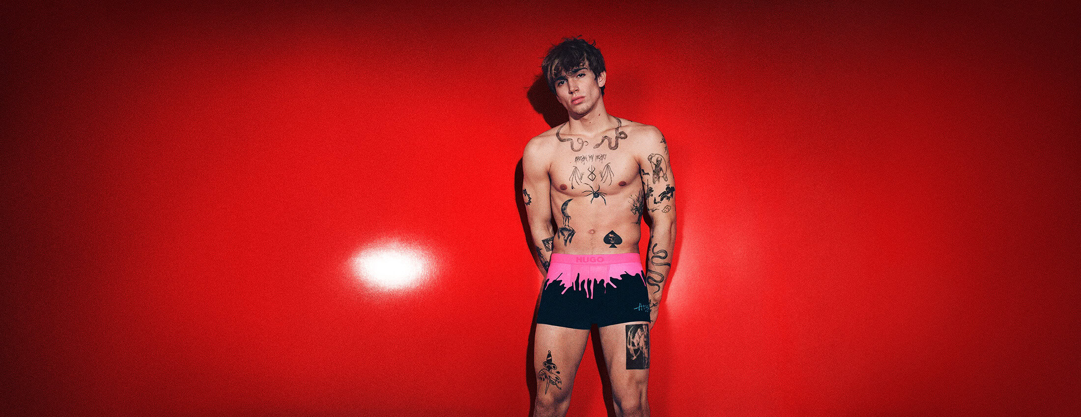 Male fashion model poses shirtless against a bright red background, showcasing torso tattoos and designer underwear in a studio setting.