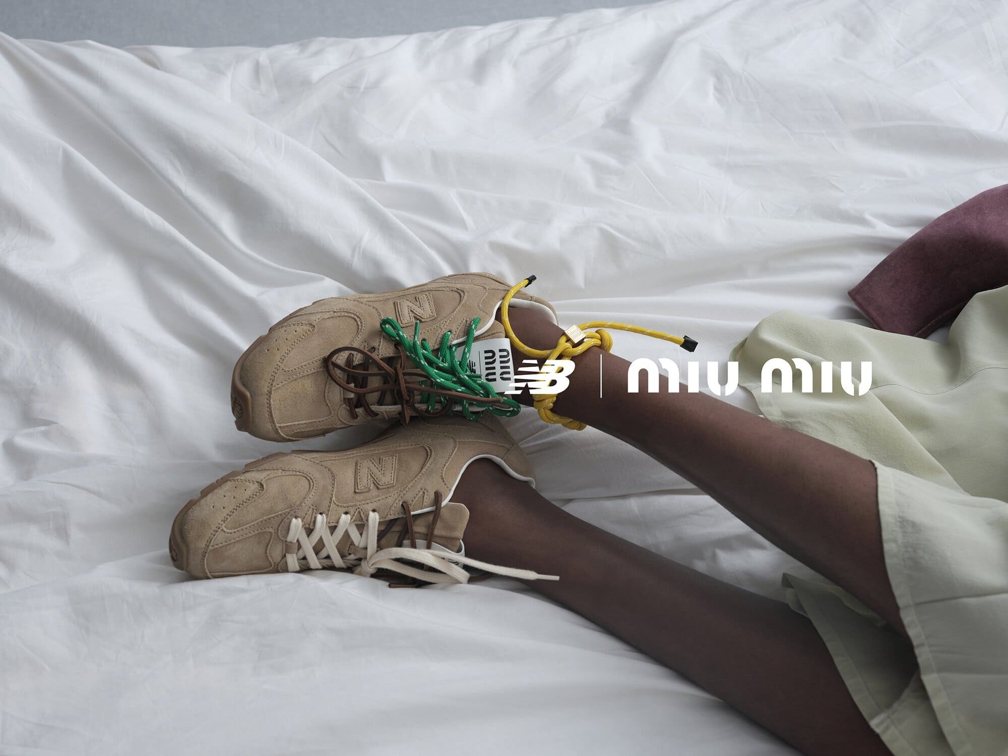 Tan New Balance sneakers are styled with a mustard yellow and green rope tie, seen on a bed with an olive-toned skirt.