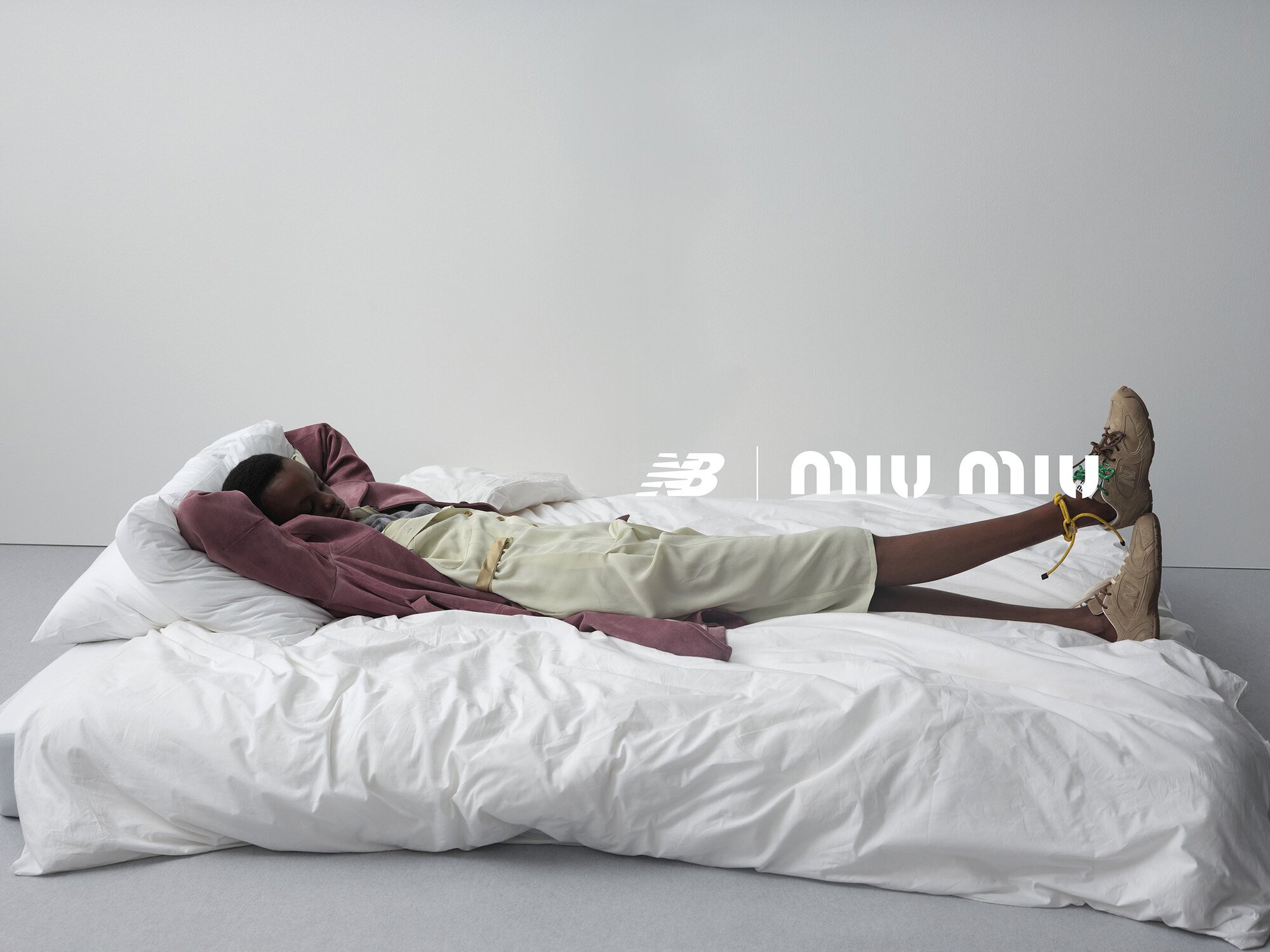 Person lies on a bed of white linens wearing athletic shoes next to the logos of New Balance and Miu Miu on a white background.