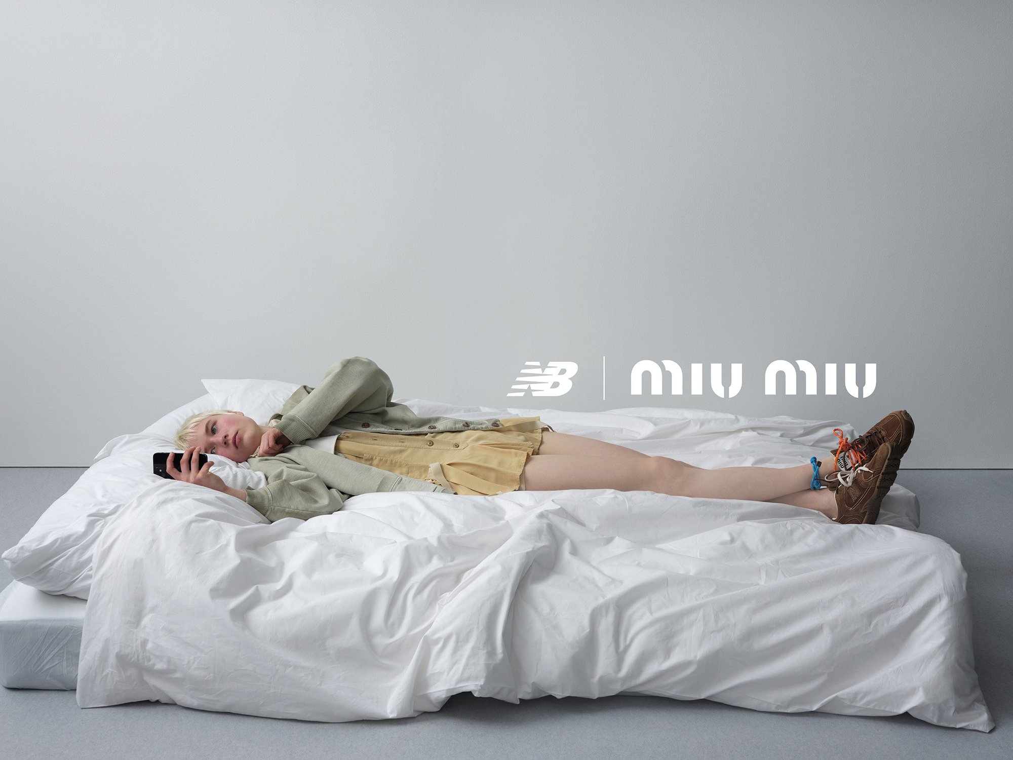 Woman lying on a white bed using a smartphone, wearing a jacket, skirt, and sneakers, against a plain gray studio background.