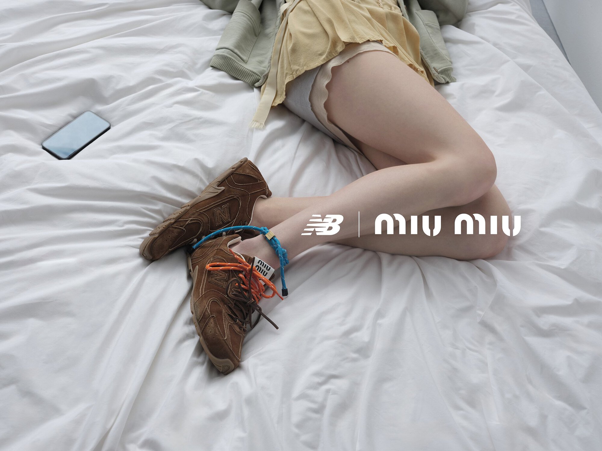 Editorial fashion shot in a bedroom. Brown sneakers on a woman's legs in bed, phone, and New Balance x Miu Miu branding.