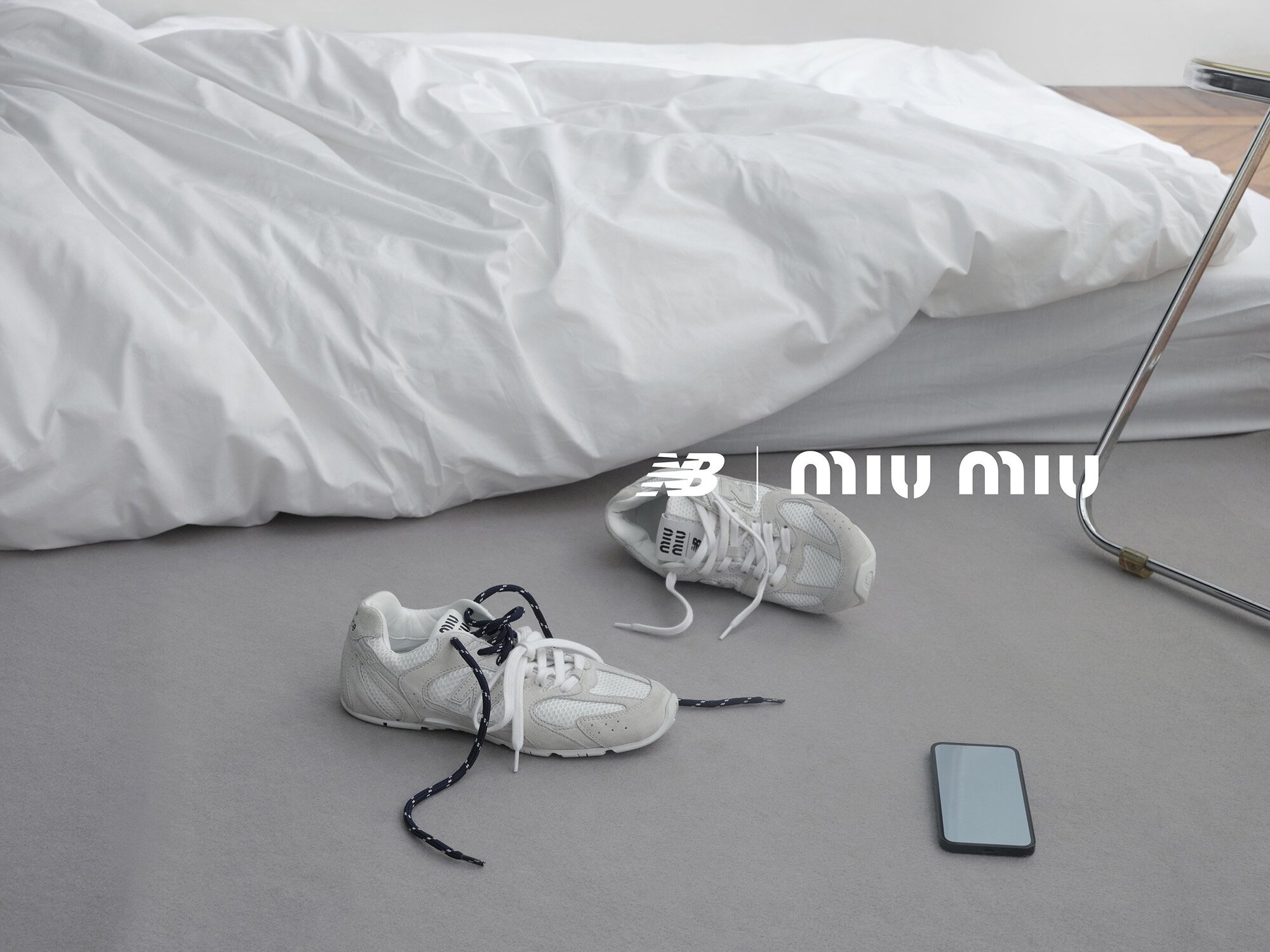 Two New Balance x Miu Miu sneakers are displayed in a minimalist setting with a mobile phone, under a white duvet cover. The scene is softly lit.