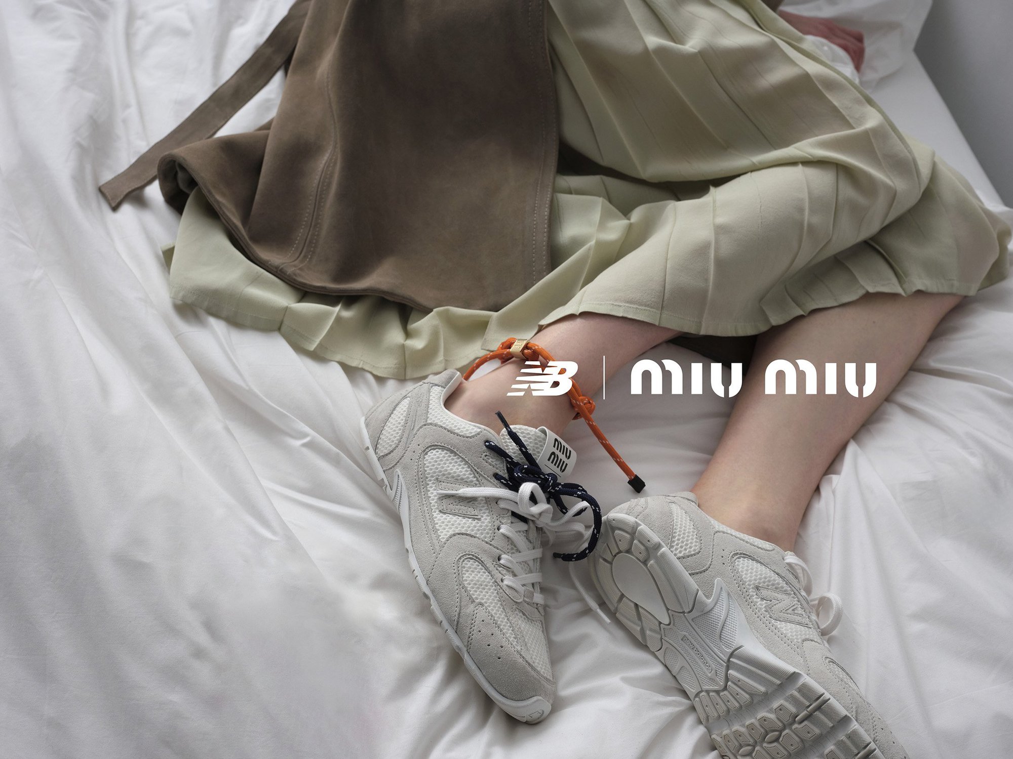 A person lies on a bed with white sheets, showcasing grey and white sneakers, a light green skirt, and a draped brown garment.