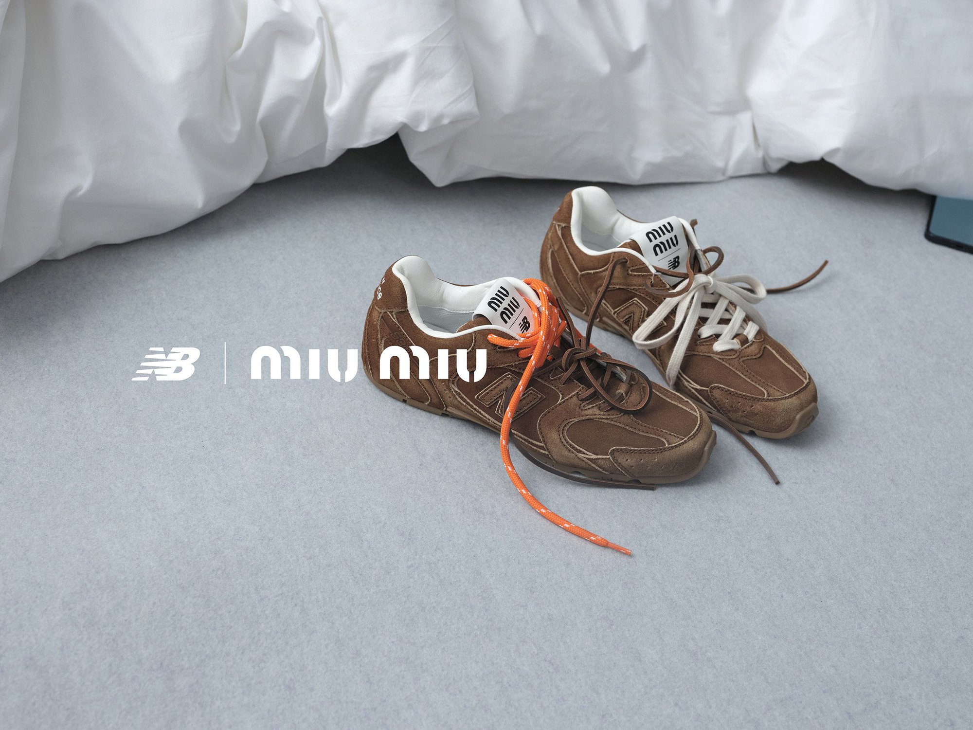 A pair of brown Miu Miu x New Balance sneakers are showcased against a grey surface with soft white bedding in the background.