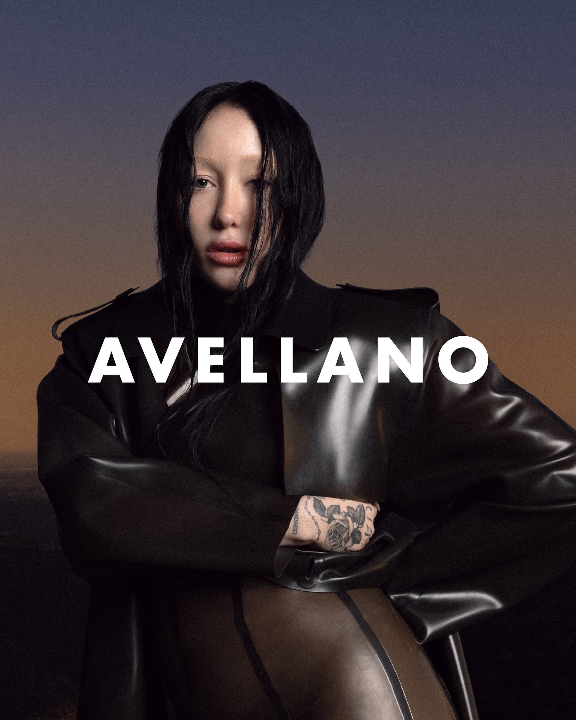 Fashion portrait of a woman in a black coat against a gradient sky background. She has dark hair and fair skin. Avellano text overlay.