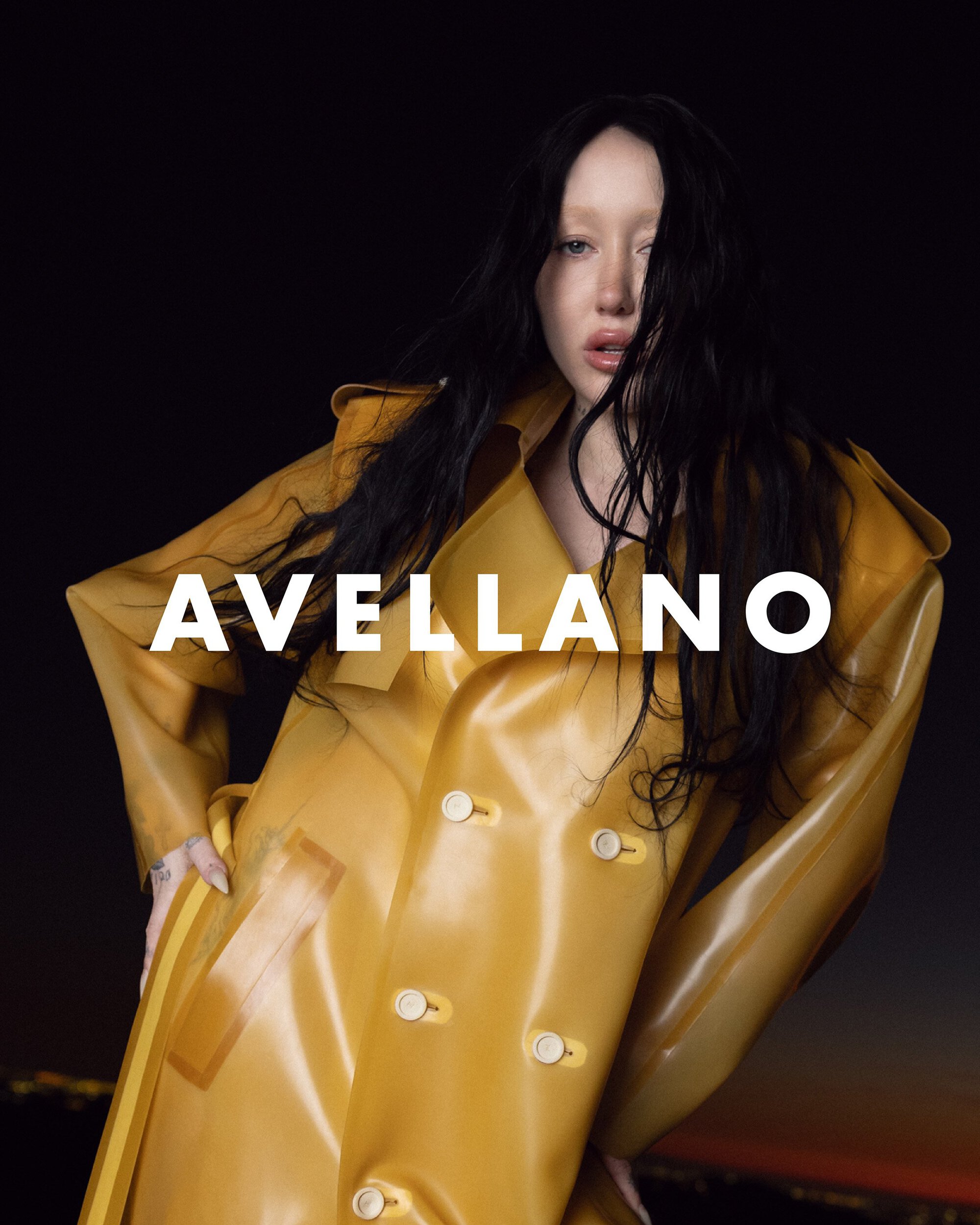 Fashion shot of a model wearing a yellow, transparent trench coat with black hair against a dark background with city lights visible.