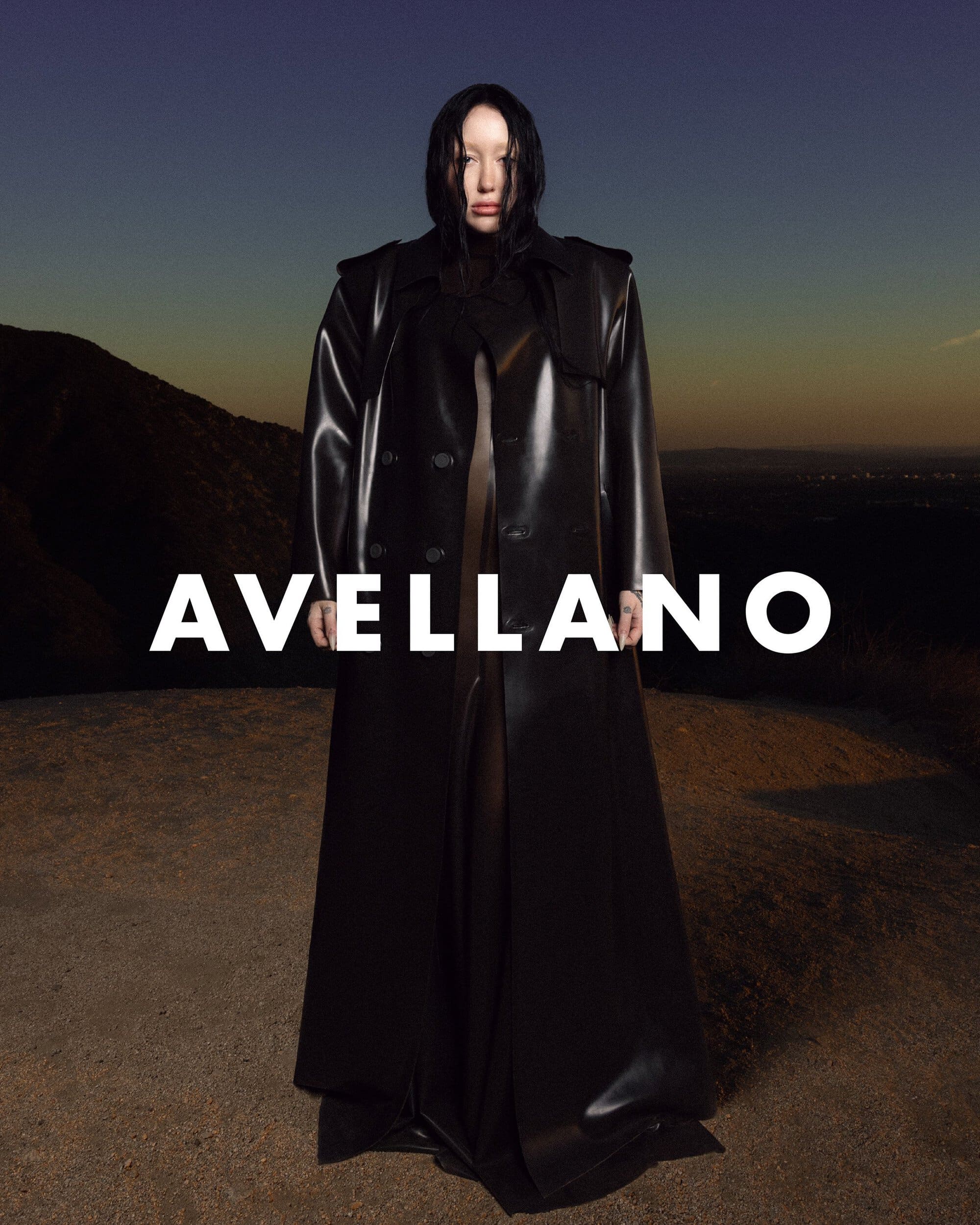 Woman in black trench coat poses against an outdoor sunset landscape. The word 'AVELLANO' is overlaid in white font.