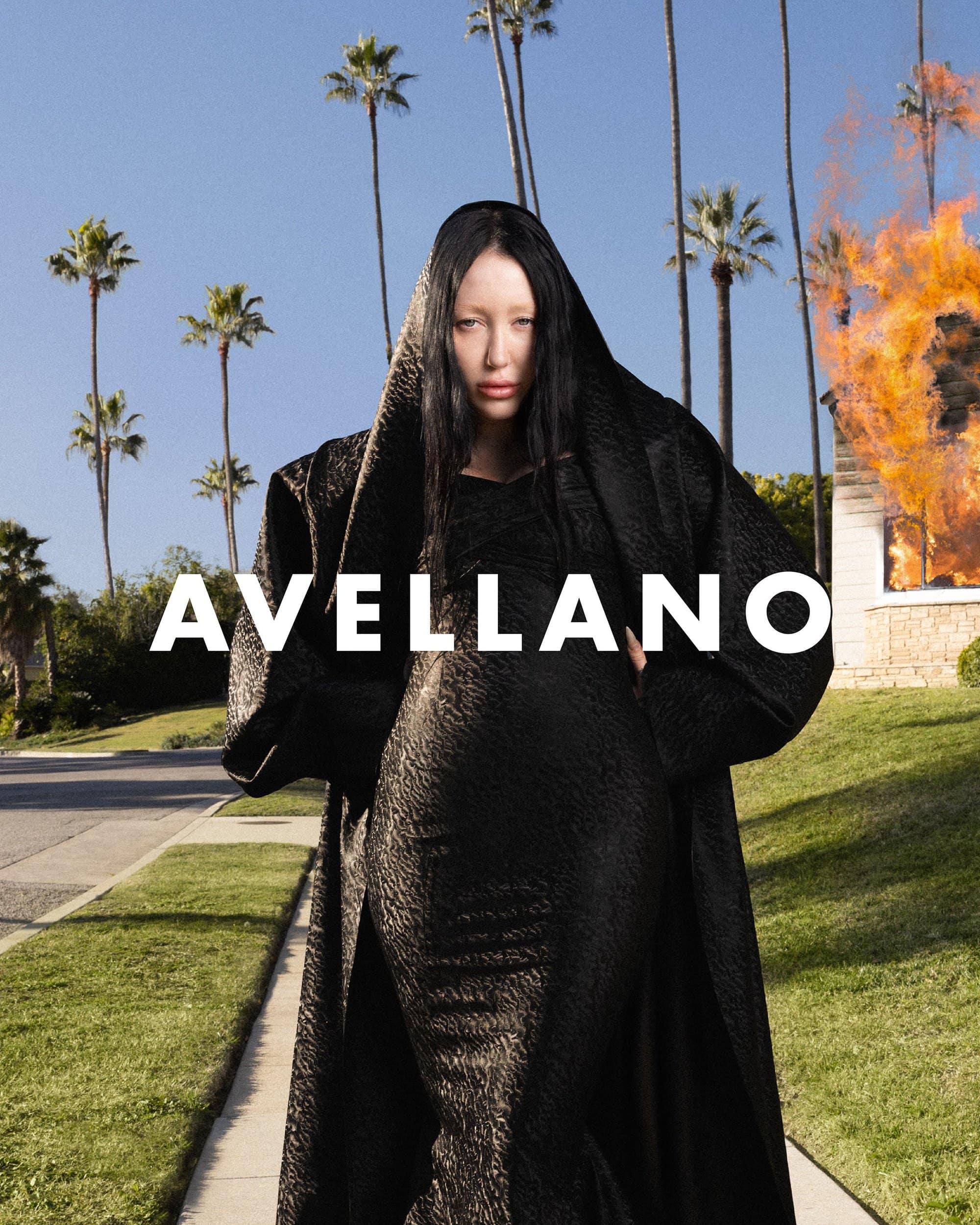 A woman wearing a long black hooded dress stands on a suburban street with palm trees, with a burning house in the background.