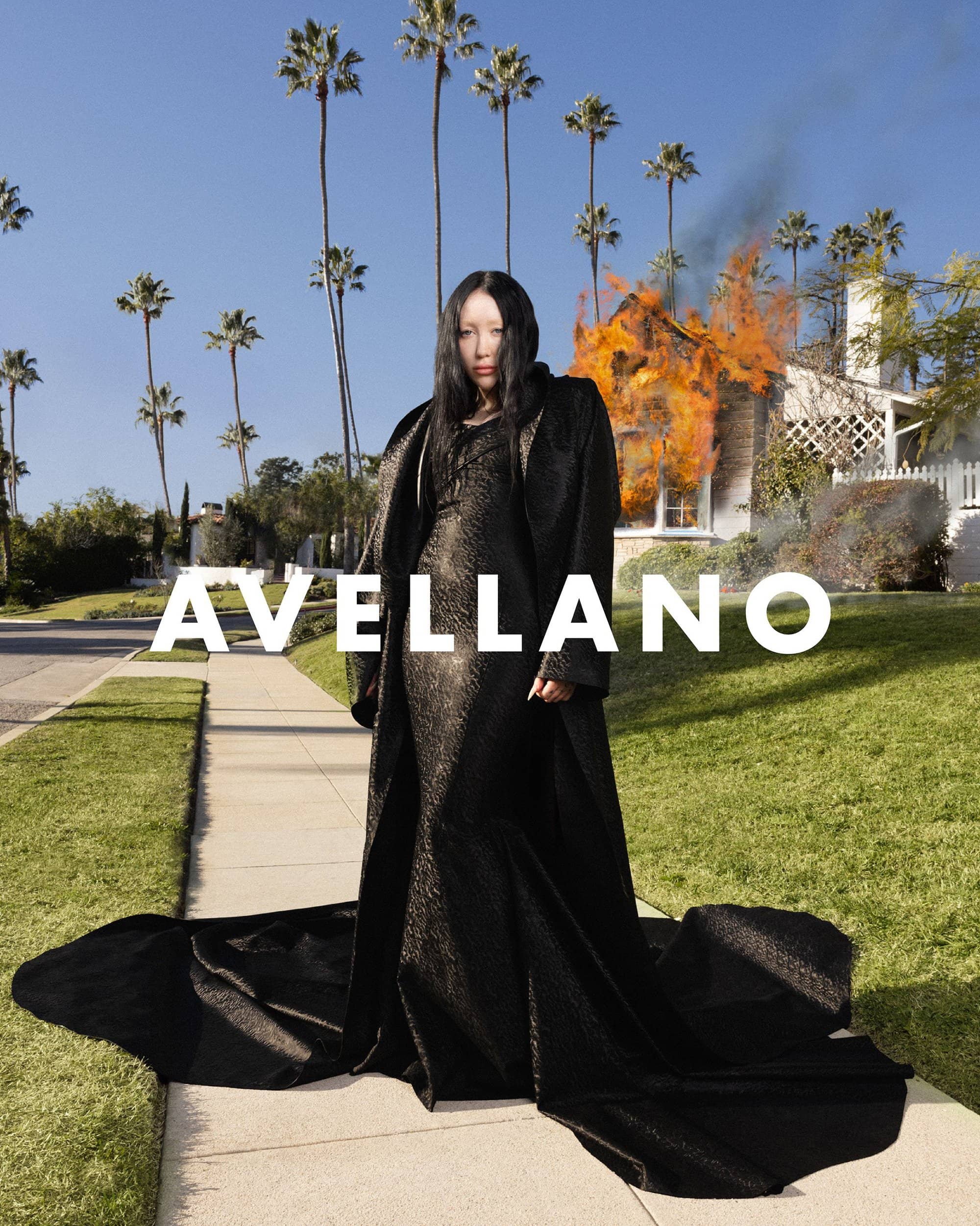 A woman in a long black coat poses on a sidewalk in front of a house on fire, set against a backdrop of palm trees and a blue sky.