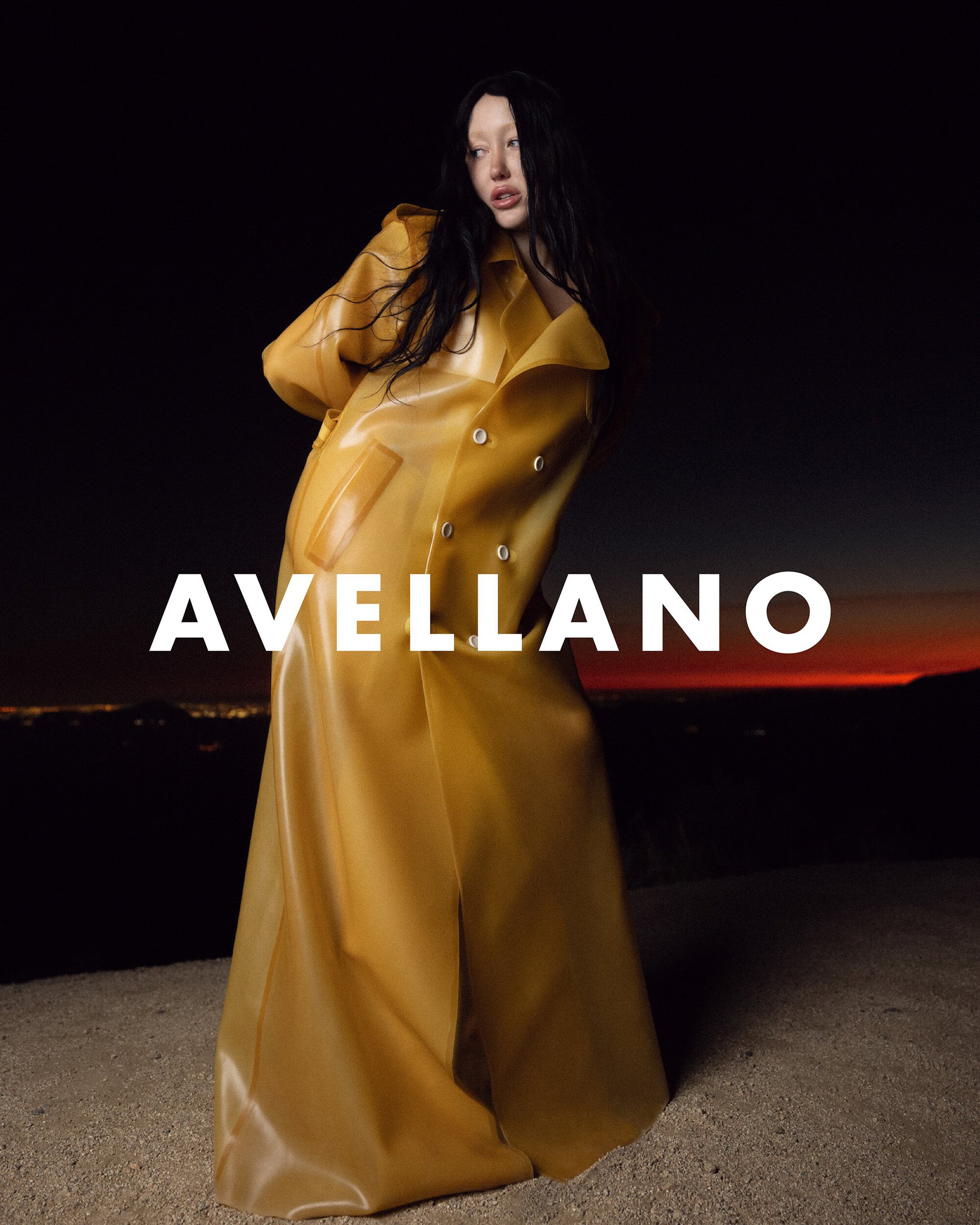 A woman in a shiny yellow coat stands in front of a dark background with a distant city skyline under a red sunset. Fashion shot.