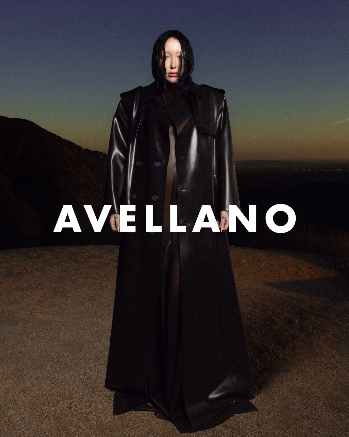 Model Noah Cyrus wearing a long black latex trench coat standing outdoors against a mountain sunset background.