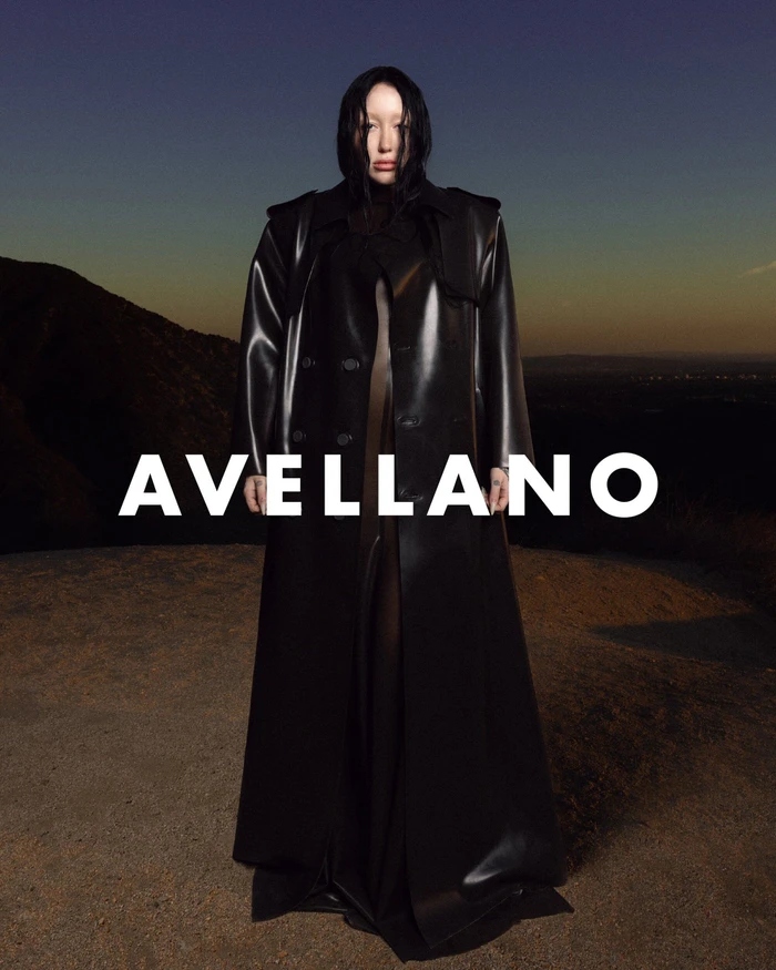 Model Noah Cyrus wearing a long black latex trench coat standing outdoors against a mountain sunset background.