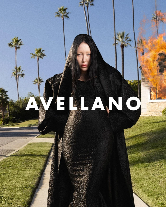 Noah Cyrus wearing a black textured hooded coat outdoors on a residential street with palm trees and a burning house.