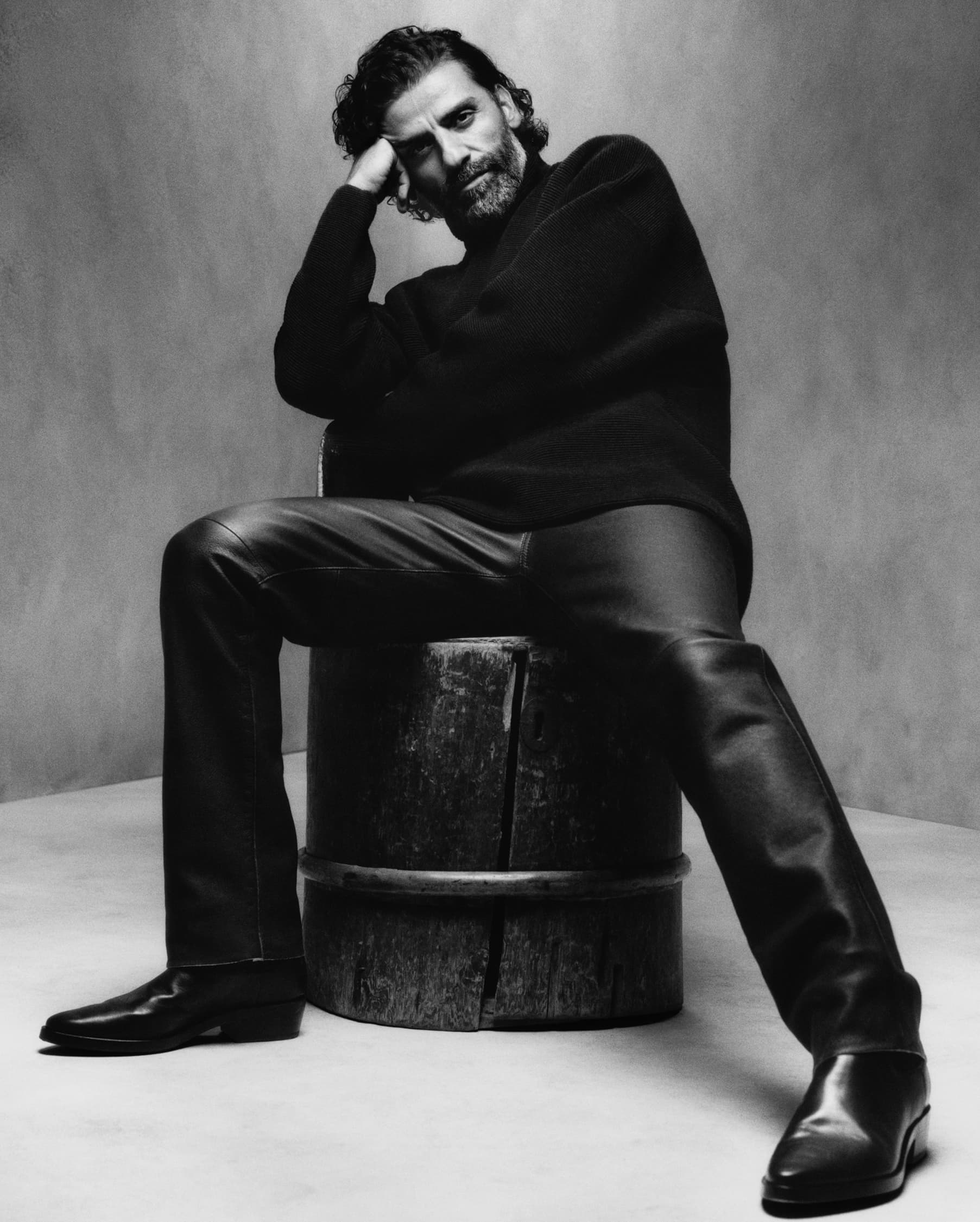 Black and white portrait of a bearded adult male sitting on a wooden barrel wearing leather pants and sweater in a photo studio.