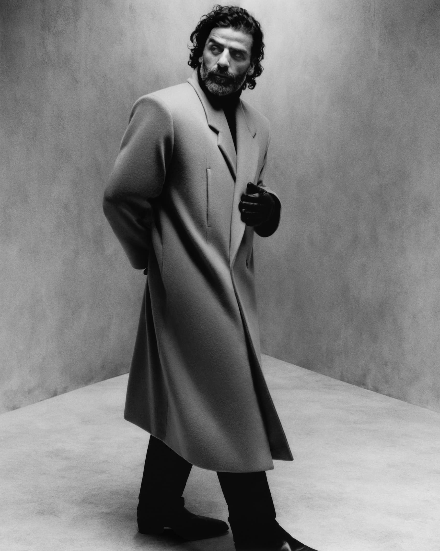 Actor Oscar Isaac poses indoors wearing an overcoat, black pants, and shoes. Soft lighting highlights his facial features.