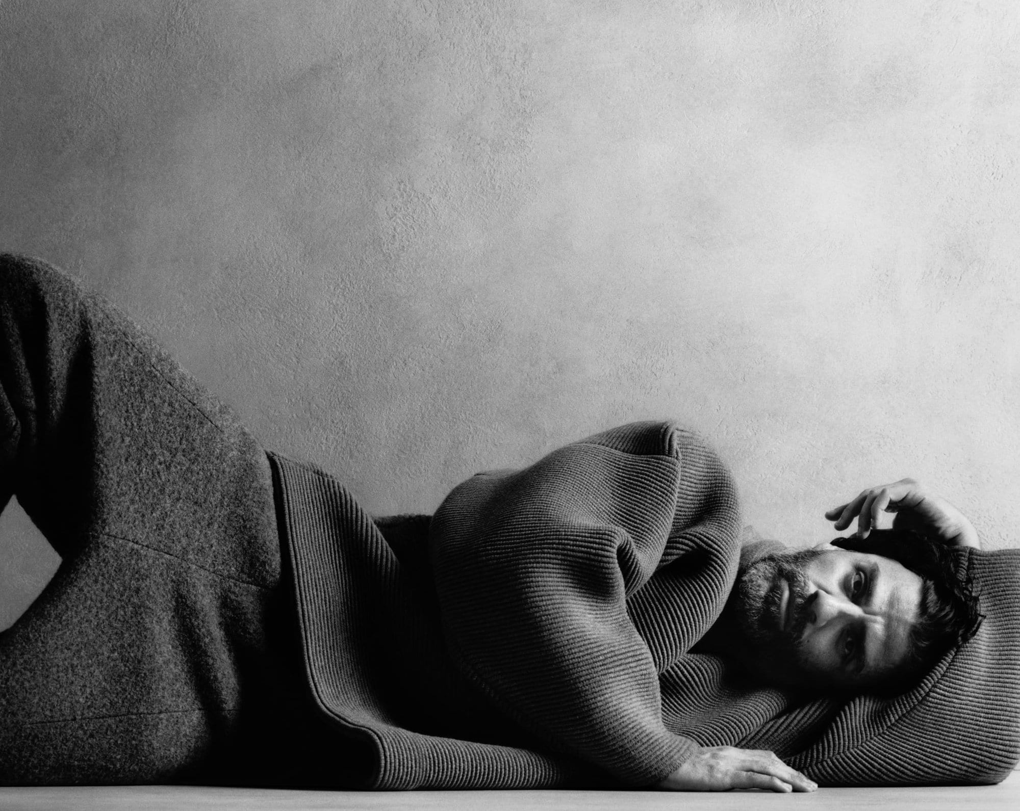 Black and white portrait of a man lying on his side wearing a sweater and pants, with a textured gray wall in the background.
