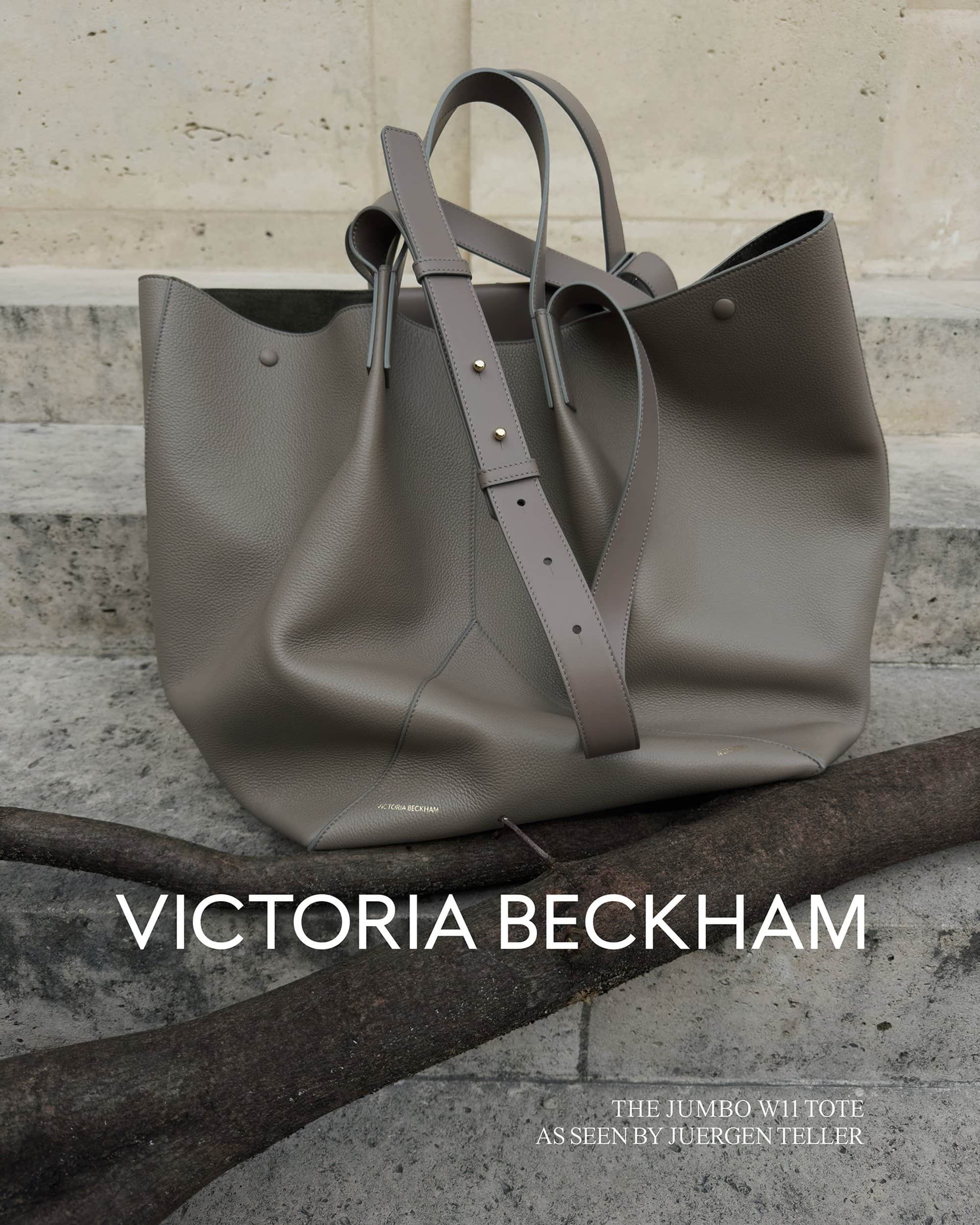Grey leather Victoria Beckham tote bag with handles and shoulder strap displayed indoors with a textured wall backdrop.