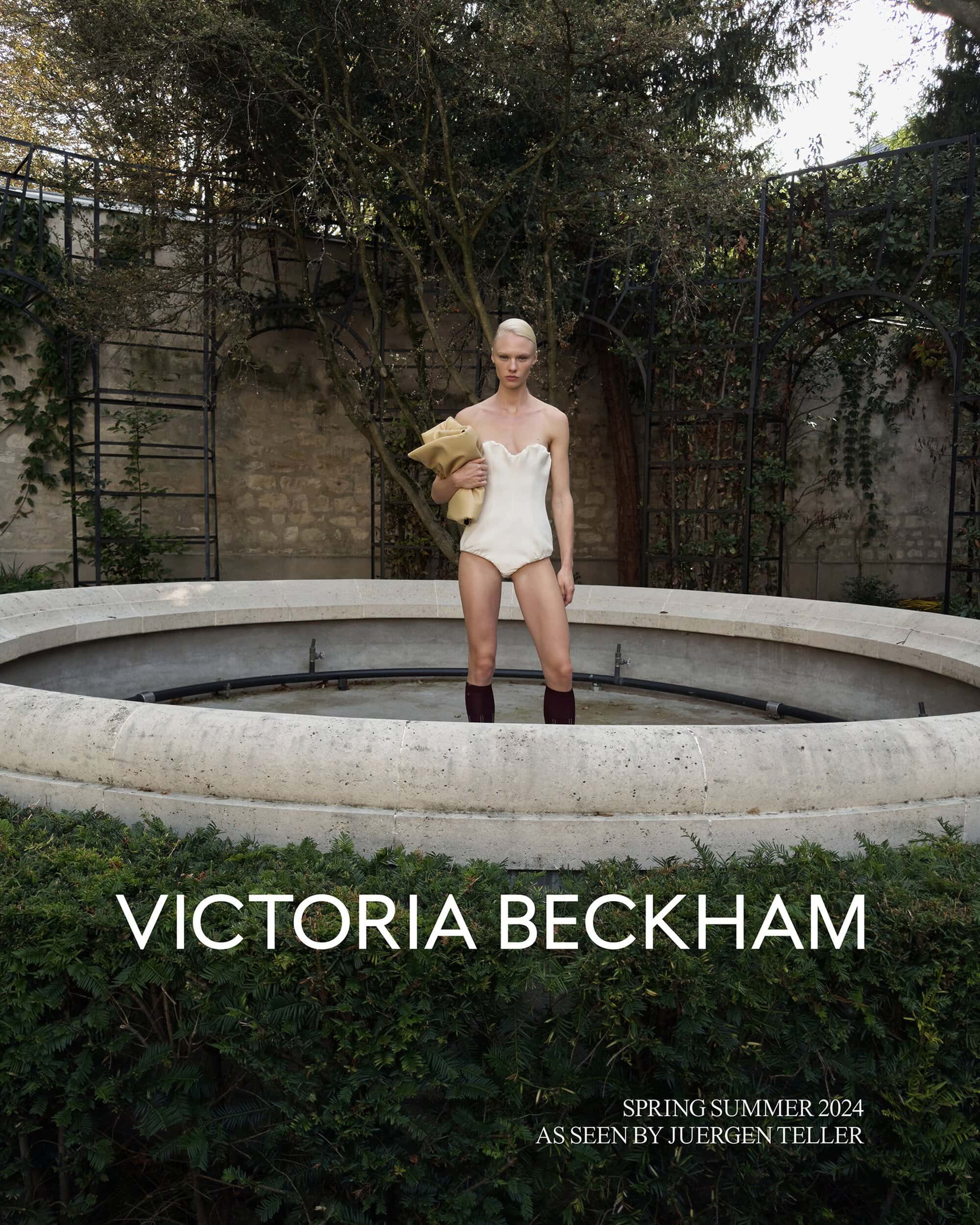 Model in cream-colored swimsuit and boots stands in an empty concrete fountain surrounded by green bushes and trees in an outdoor setting.
