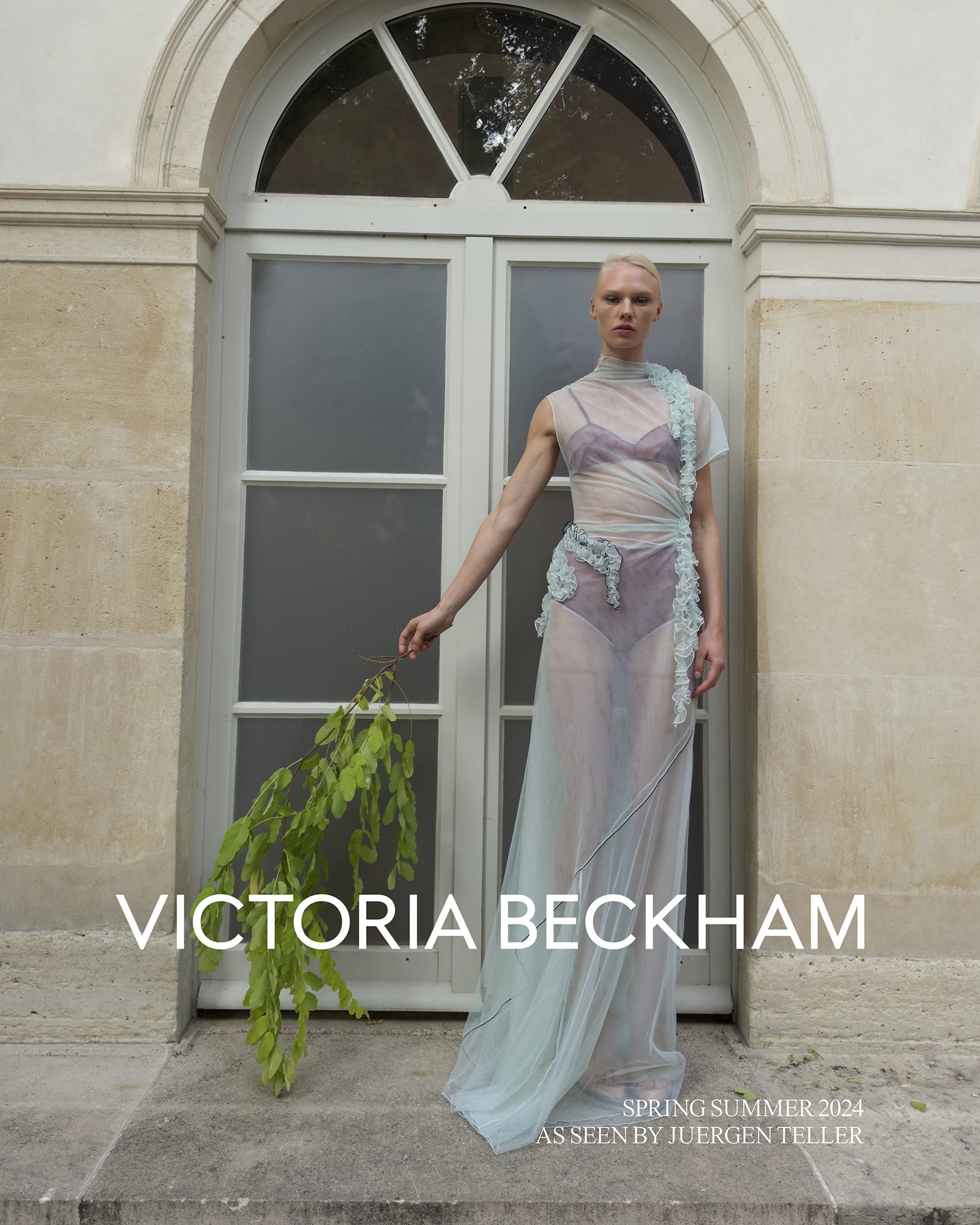 A slender adult fashion model poses outdoors in front of white-painted doors wearing a translucent light blue dress with floral trim.