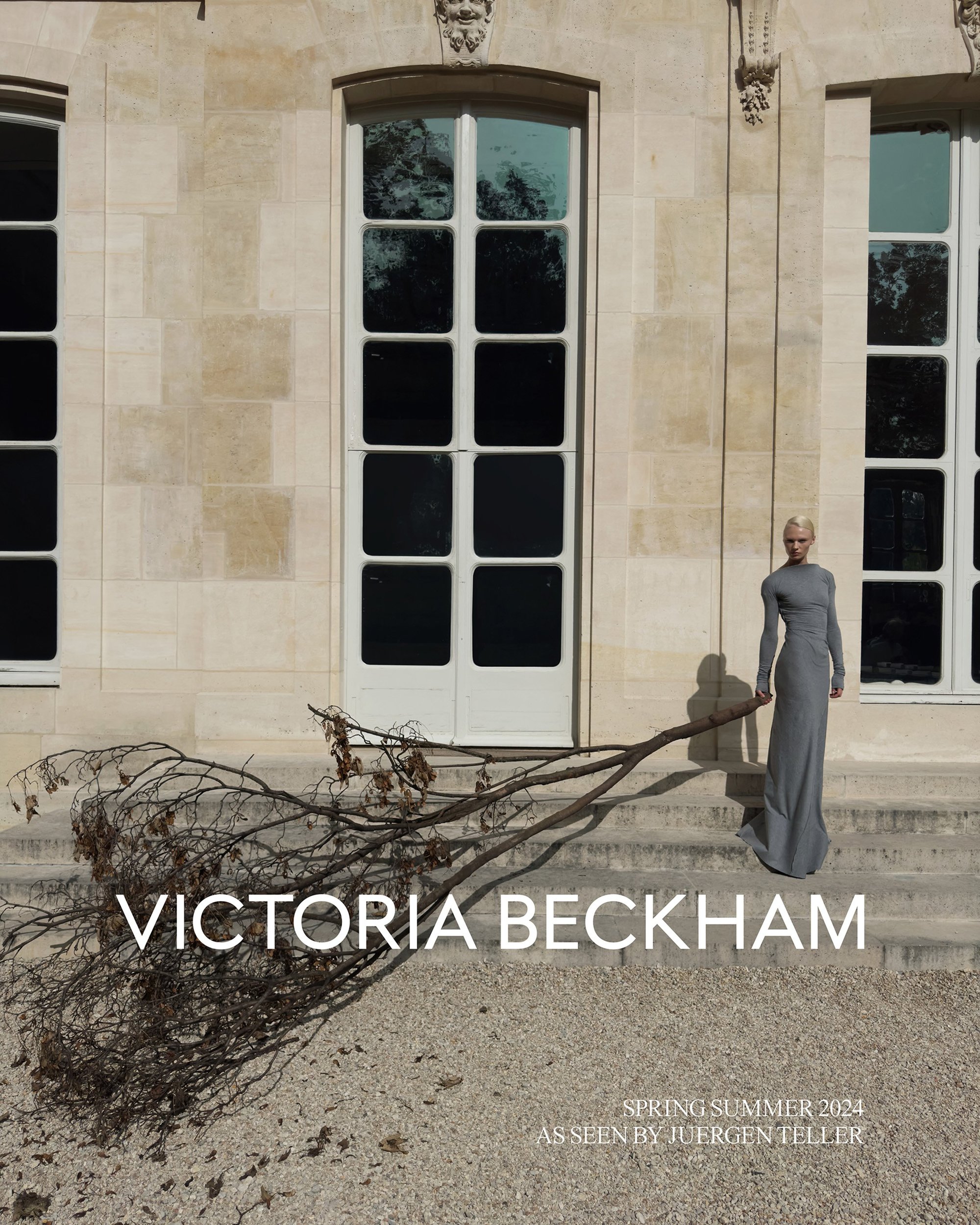 Victoria Beckham stands on stone steps of a building holding a large bare branch wearing a long grey gown for a fashion ad.