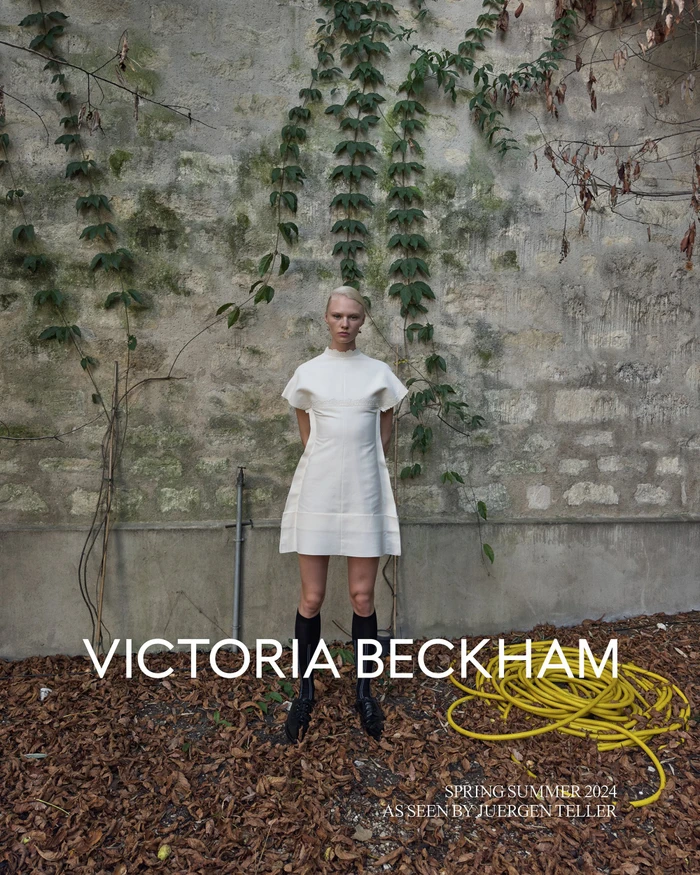 Model wearing a white minidress and black knee-high socks standing before a stone wall covered in vines for Victoria Beckham.