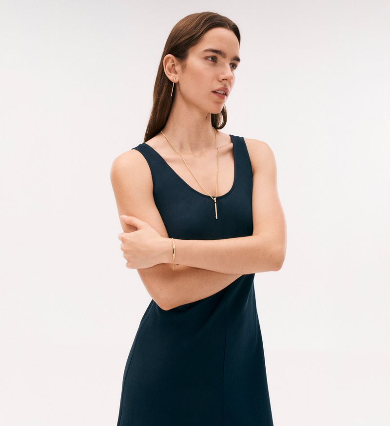 A woman in a navy dress stands against a white background, accessorized with gold jewelry, her arms crossed in a studio setting.