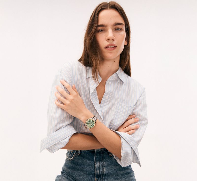 An adult woman poses indoors against a bright background, wearing a striped shirt and denim jeans with a wristwatch on her left wrist.