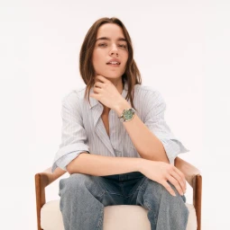 A woman wearing a striped shirt and blue jeans sits in a chair, displaying a wrist watch against a white background.