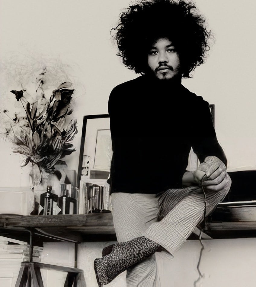Monochromatic portrait of an adult male with a large afro hairstyle, wearing a black turtleneck and patterned pants, seated indoors.