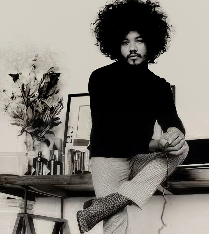 Man with voluminous afro hair wearing a black turtleneck and patterned trousers leaning against a desk in a monochrome studio setting.