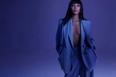 Naomi Campbell wearing an oversized blue suit jacket and trousers against a dark blue background.