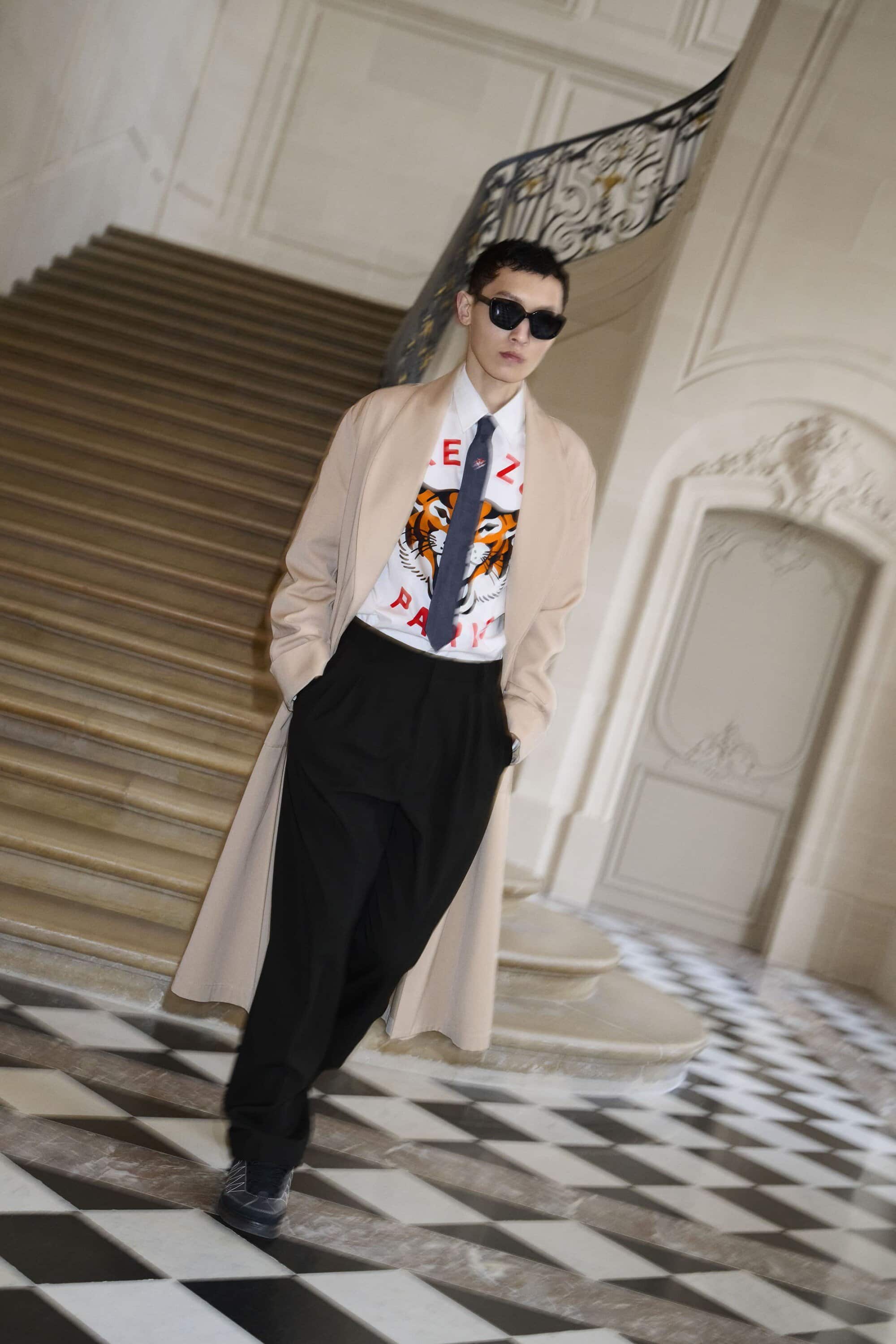 Stylish adult male in a beige trench coat, graphic tee, and dress pants poses indoors with a black and white checkered floor.