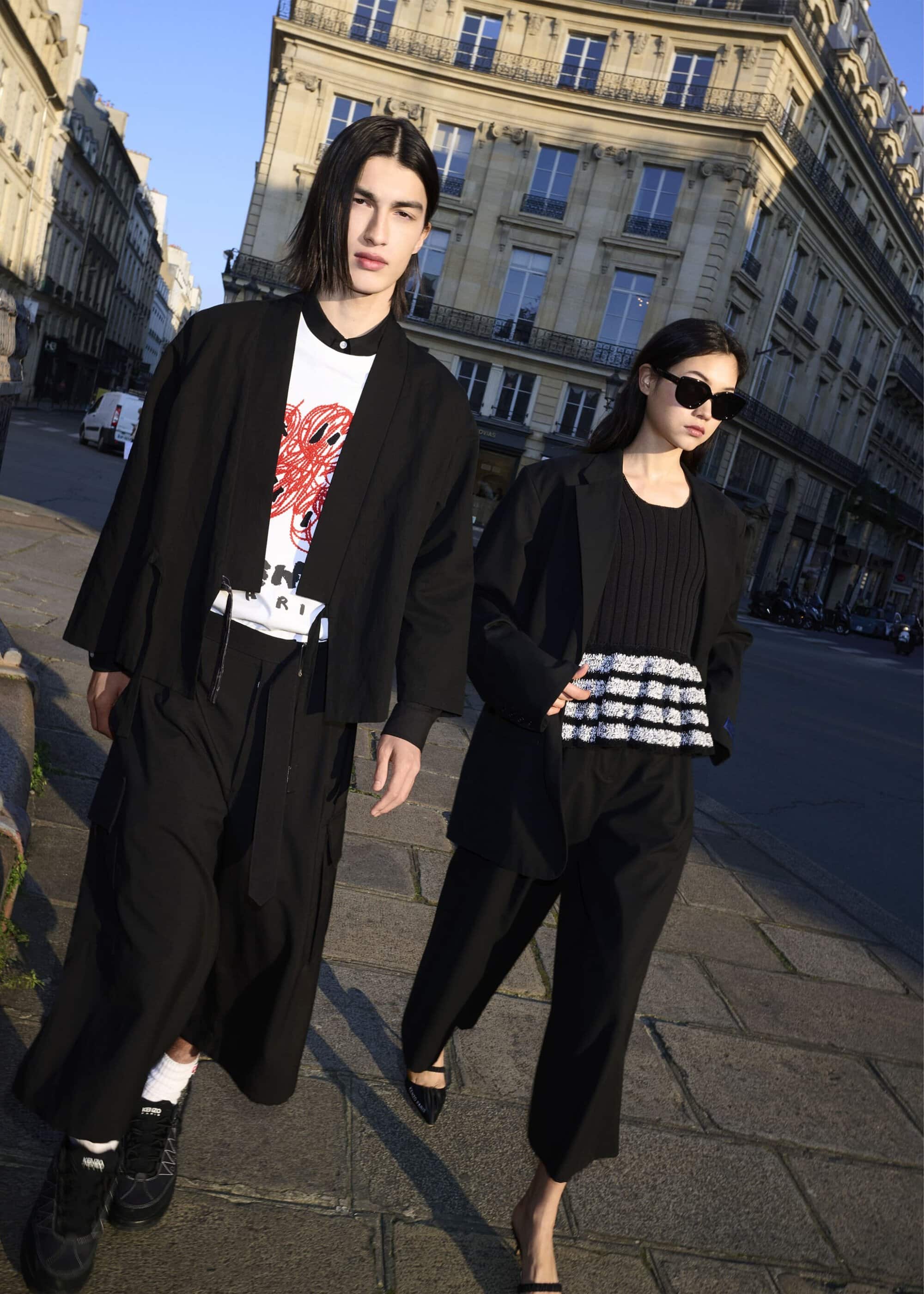 Two fashion models pose in an urban street setting, wearing black and white outfits with modern silhouettes, showcasing contemporary fashion.
