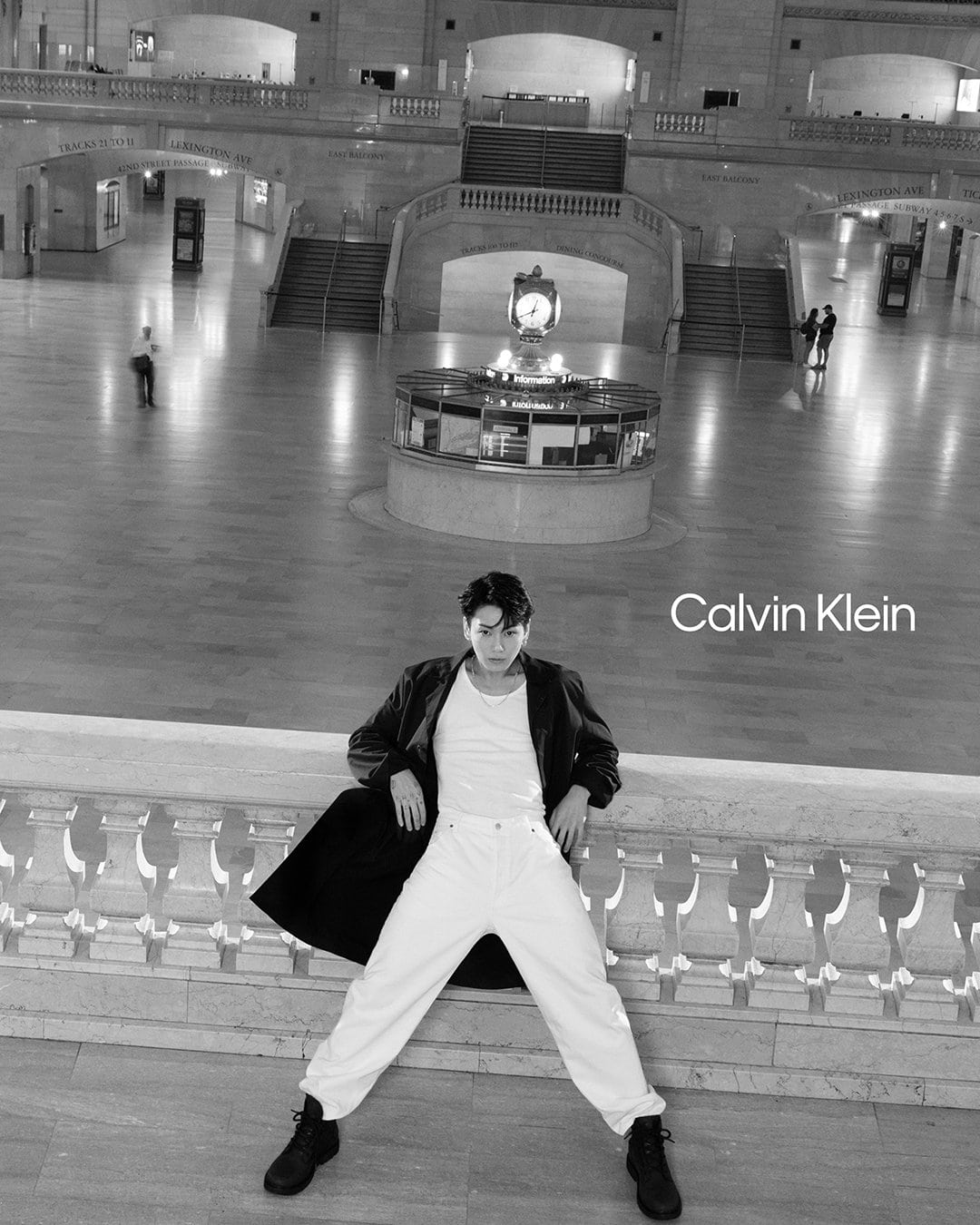 Monochrome image of a male model leaning against a balustrade in a grand hall wearing white jeans, a white shirt, and a dark jacket.