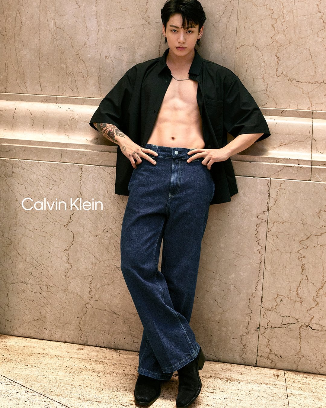 Male model poses in open shirt and jeans with a marble wall background, highlighting fashion and physique.