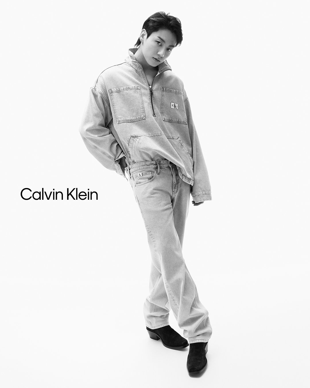 Black and white full-body shot of a young adult male fashion model in denim jacket and jeans, posing on a white backdrop in a photo studio.