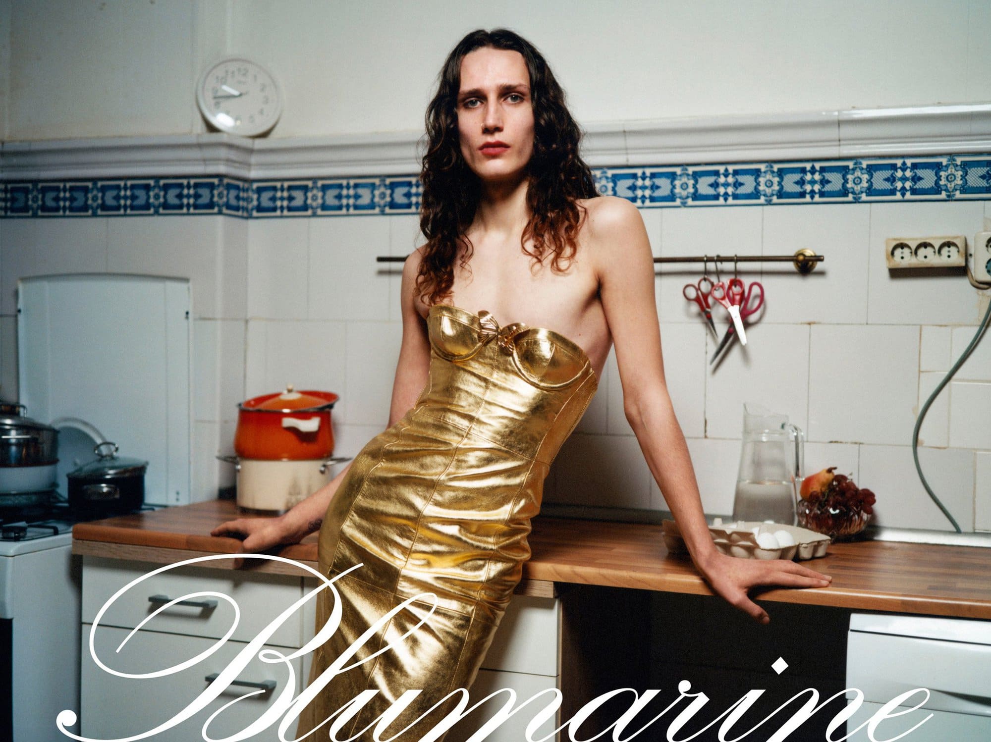 A woman in a gold dress poses in a kitchen with white tiled walls and retro appliances, advertising Blumarine fashion.