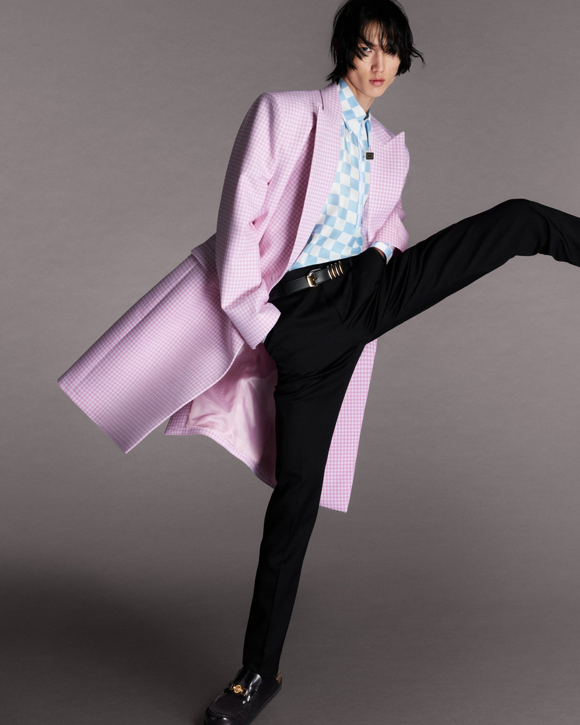 Fashionable young adult with pink and white checkered coat poses in a photo studio wearing black pants and shoes.