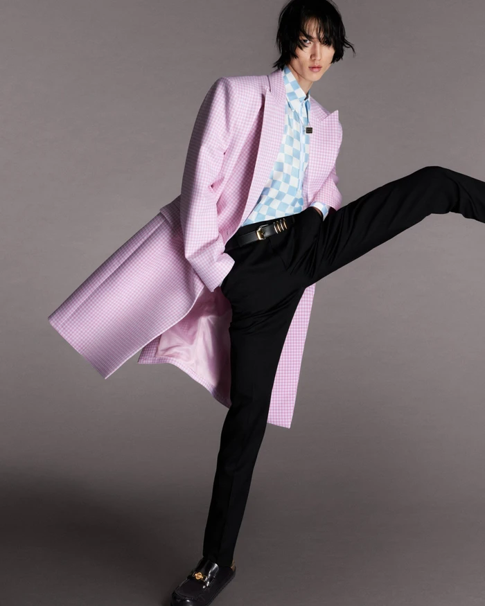 Man wearing a light pink houndstooth coat, checkered shirt, and black trousers against a neutral grey studio background