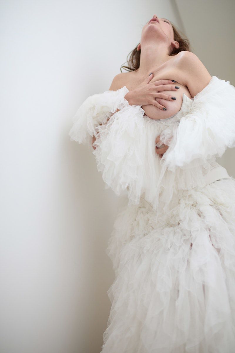 Adult female poses in a flowing white ruffled dress against a plain wall, casting shadows and capturing an editorial aesthetic.