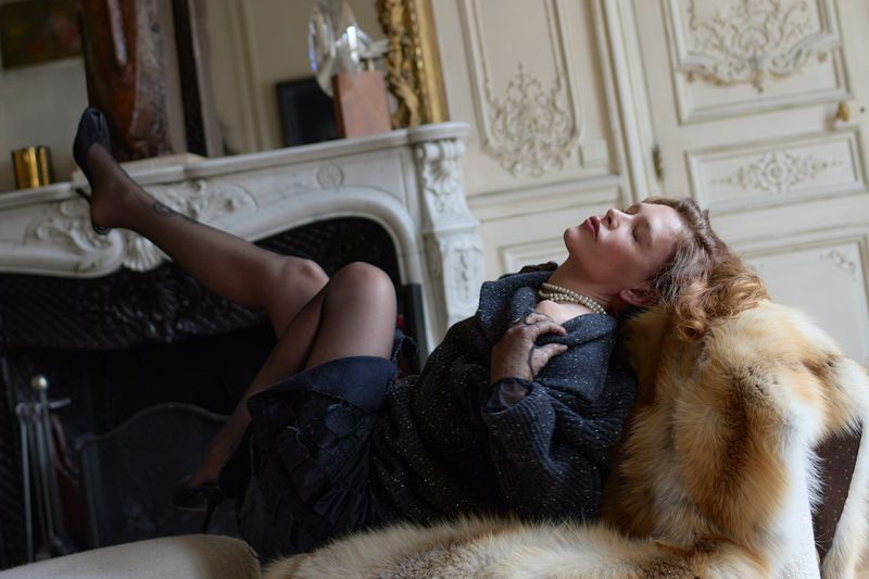 Glamorous woman in black dress and stockings reclining on a fur throw in a vintage-style room with a fireplace and ornate walls.